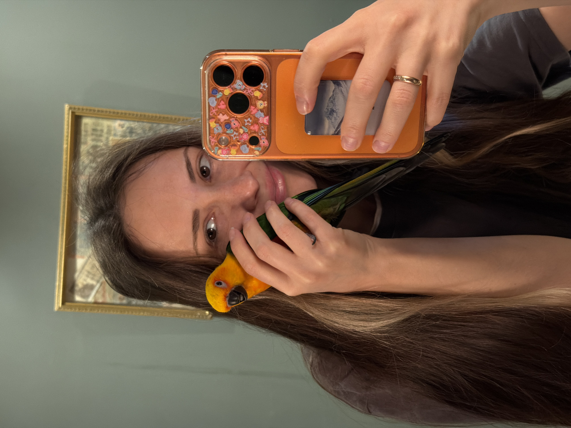 A woman is taking a selfie in a mirror with a yellow and green parrot. She is holding an orange phone case with a credit card in the back. She has long brown hair with blonde highlights. There is a framed picture behind her. The background is a light green wall. There is no evidence that the image was taken in Khimki, Russia.