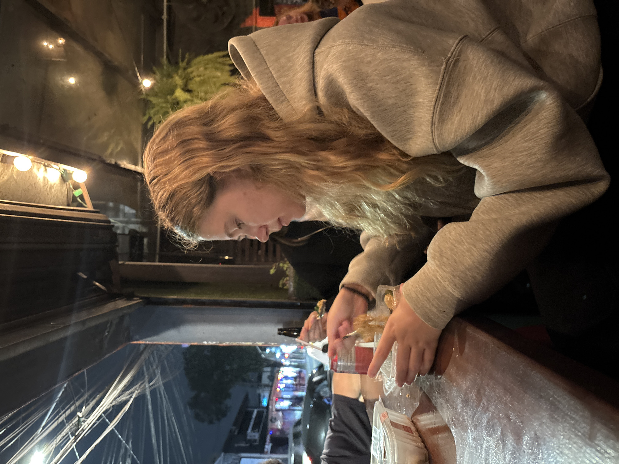 A young woman with long, wavy blonde hair and wearing a grey hooded sweatshirt leans over a table. She appears to be handling food in clear plastic containers. The scene takes place outdoors at night. There are lights overhead and a glimpse of a street with colorful lights, potentially a commercial area. Electrical wires can be seen overhead. Given the urban setting and the presence of streetlights and commercial establishments, it is possible the location is in São Paulo, Brazil. Further information is needed to confirm.