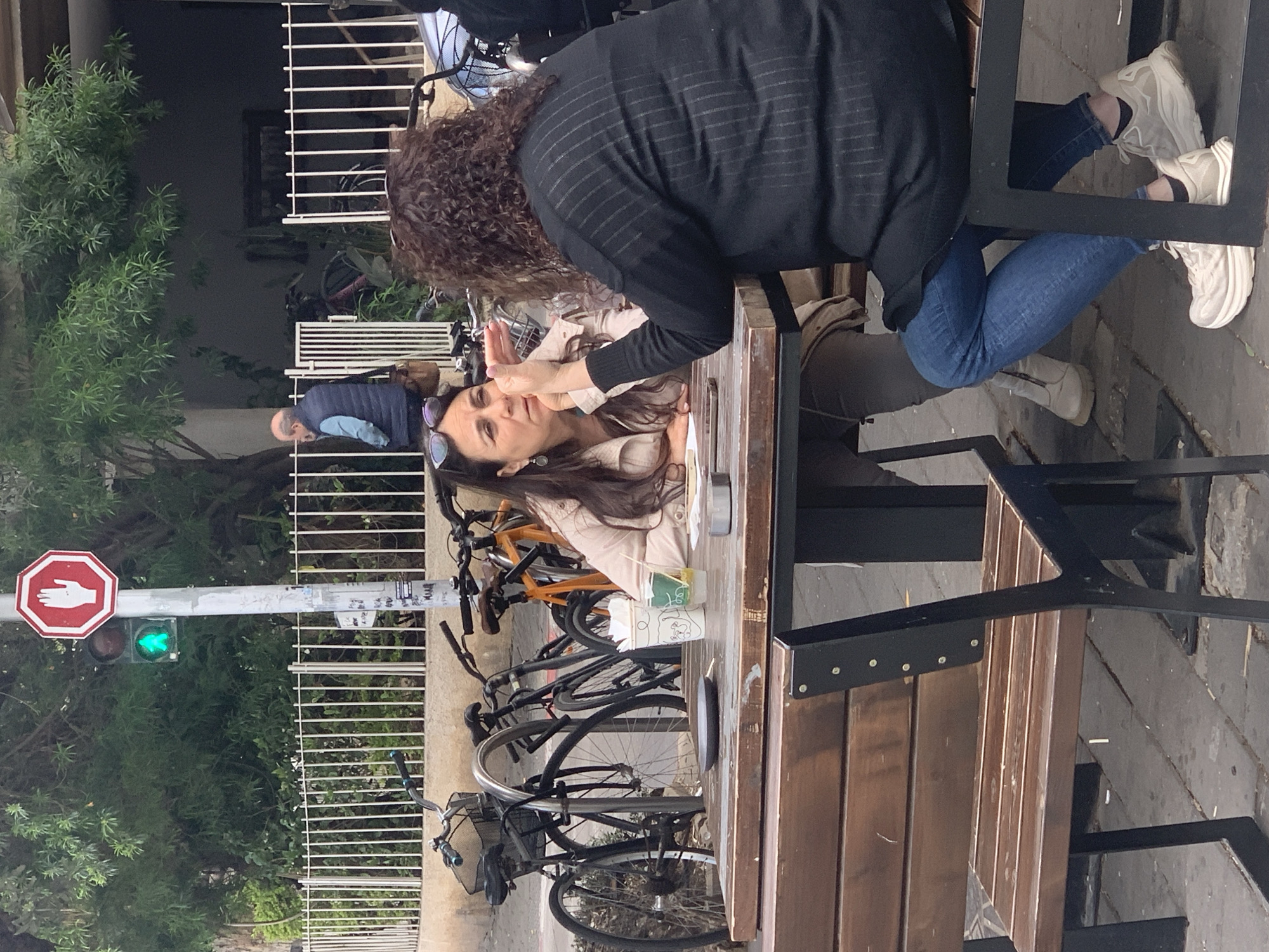 The image captures an outdoor scene in Tel Aviv, Israel. Two women are seated at a wooden table, engaged in conversation. One woman, facing the camera, has brown hair and wears sunglasses on her head. She has her hands raised near her face. The other woman has curly brown hair and is wearing a black striped top. A man in a blue vest is visible in the background.
Several bicycles are parked next to the table. To the left of the frame, a street sign with a red octagon with a white hand is mounted on a pole, under which a pedestrian crosswalk signal with a green walking man is lit. Lush greenery fills the background.