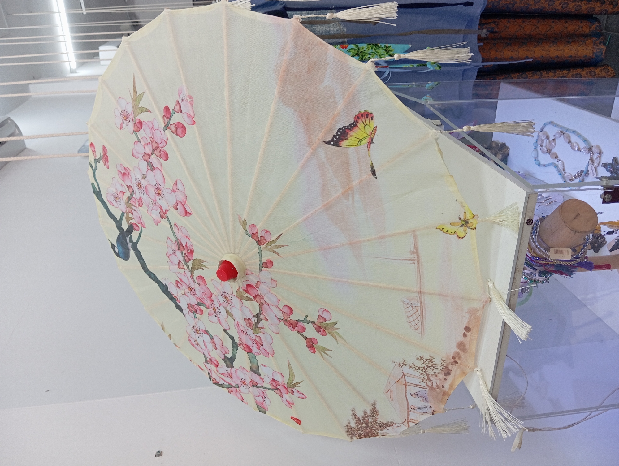 A traditional Asian umbrella, possibly silk, is displayed on a white surface against a white wall. The umbrella is open and tilted, revealing a pale yellow fabric decorated with a painted scene. The scene features a branch with pink blossoms and a bird, a watercolor landscape with a pagoda-like structure on a sandy shore with two small figures, a boat on the water, and two butterflies. The umbrella has a red knob at its apex and is adorned with white tassels hanging from its edges. Below the umbrella, a glass display case contains various beaded necklaces and other decorative items. In the background, hanging clothes and more display items are visible. The scene appears to be an indoor retail setting, possibly a shop in Nha Trang, Vietnam, specializing in artisanal goods. There are no people clearly depicted in the image.