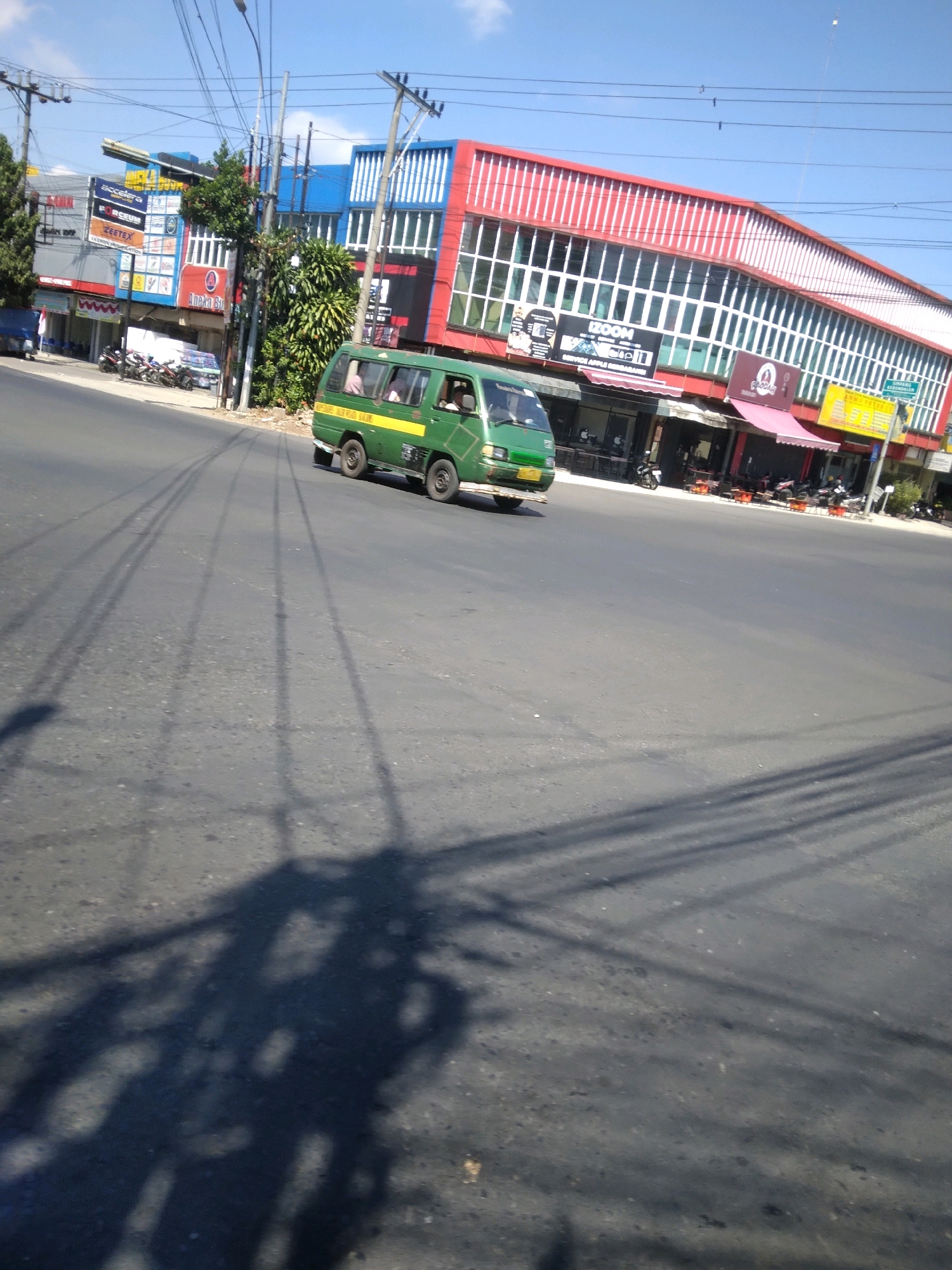 In the city of Purwokerto, Indonesia, on a bright, clear day, a green minibus drives down a wide, asphalt road. The minibus, a common mode of public transportation, is moving from left to right in the frame. The road appears to be well-maintained, with tire marks visible, indicating recent activity. The background showcases a vibrant commercial area. A multi-story building with a red and blue facade dominates the right side of the image. The building features large windows and numerous signs advertising various businesses, including "ZOOM" and "ANAKA BIRU." Several awnings are visible, some pink and some yellow, further adding to the bustling atmosphere. In front of the building, parked motorcycles suggest a lively commercial hub. On the left side of the road, a less ornate building with multiple advertisements is also present. Electrical poles and wires crisscross the sky, a typical urban feature. The sun casts distinct shadows of the electrical wires and the minibus onto the road, creating a graphic pattern across the asphalt. A lone person can be seen inside the green minibus, through the window. The overall scene depicts a typical day in a busy urban environment in Purwokerto, Indonesia.