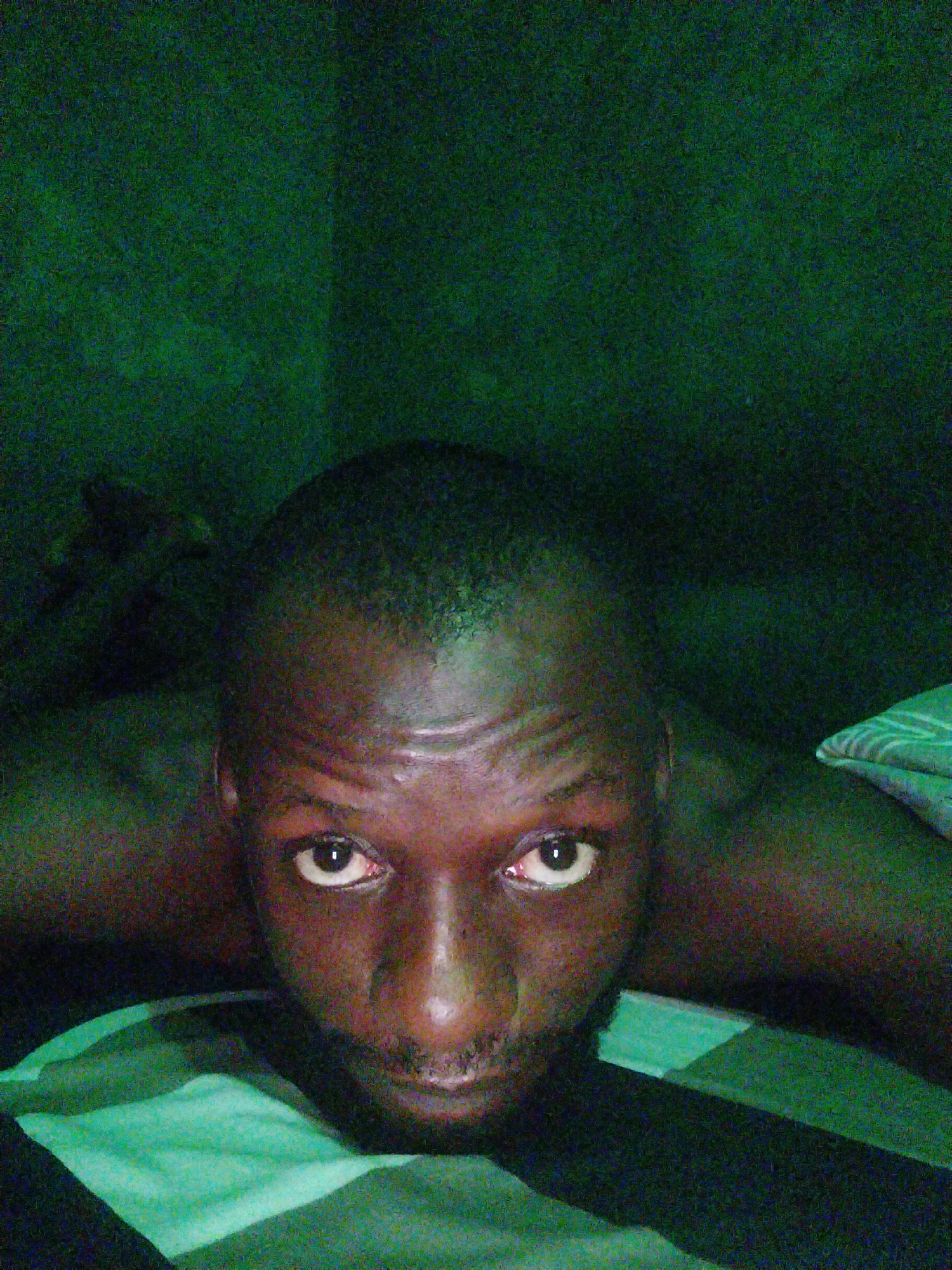 The image is a close-up of a dark-skinned man's face. He has a shaved head and a short beard. His eyes are wide open and directed at the camera. The background is dark and appears to be a wall with a green hue. He appears to be lying on a bed with a green and black striped sheet. The location and city are unknown.