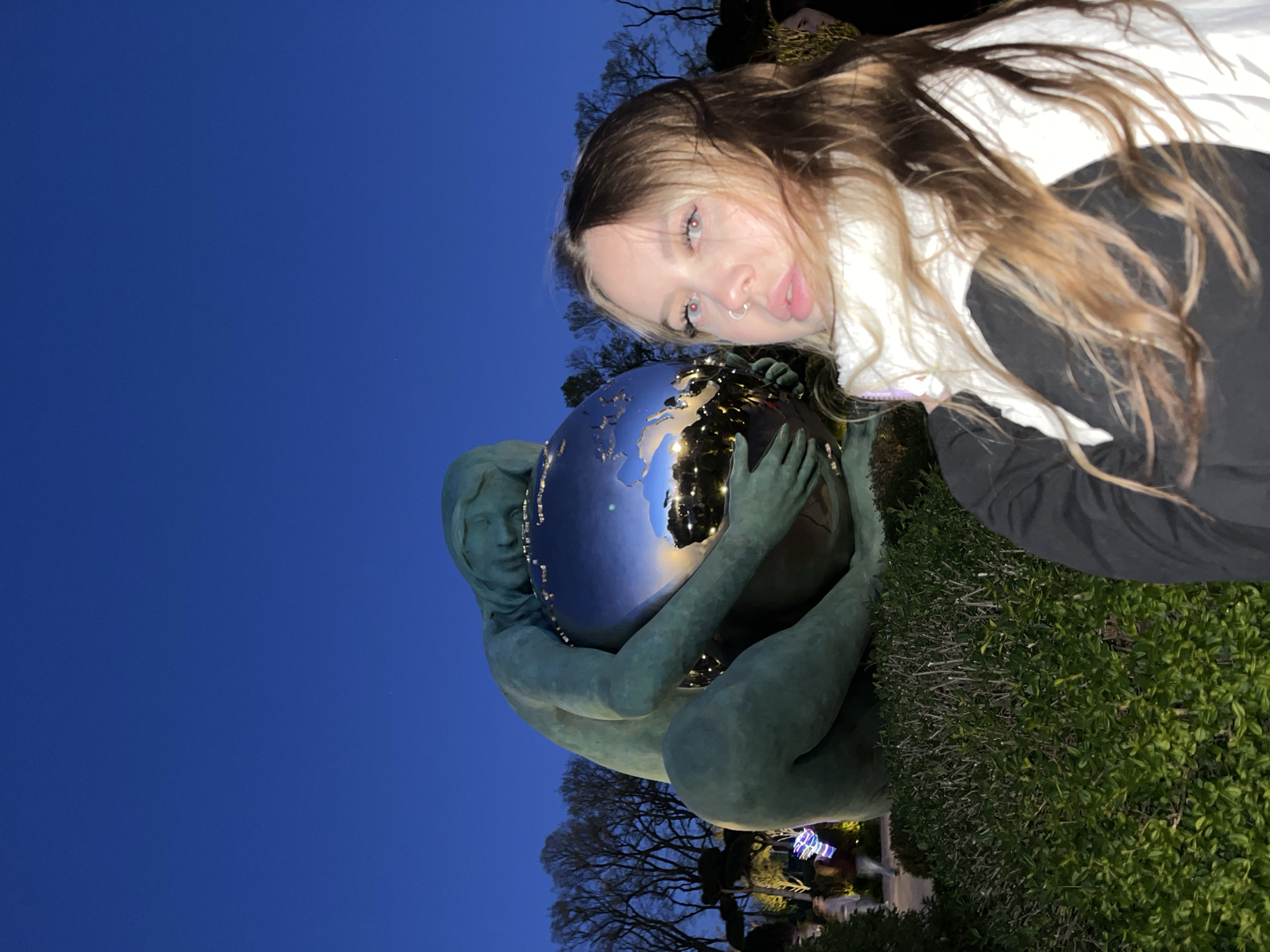A selfie taken at night shows a person with long brown and blonde hair, light eyes, and a nose ring standing beside the "Earth Keeper" statue in Krasnodar, Russia. The statue, made of a greenish material, depicts a woman holding a globe that reflects the surrounding city lights. The background features a dark blue sky, bare trees, and glimpses of other lights. The person is wearing a white top under a dark jacket. The camera angle is slightly tilted. The bottom right of the image shows green shrubbery.