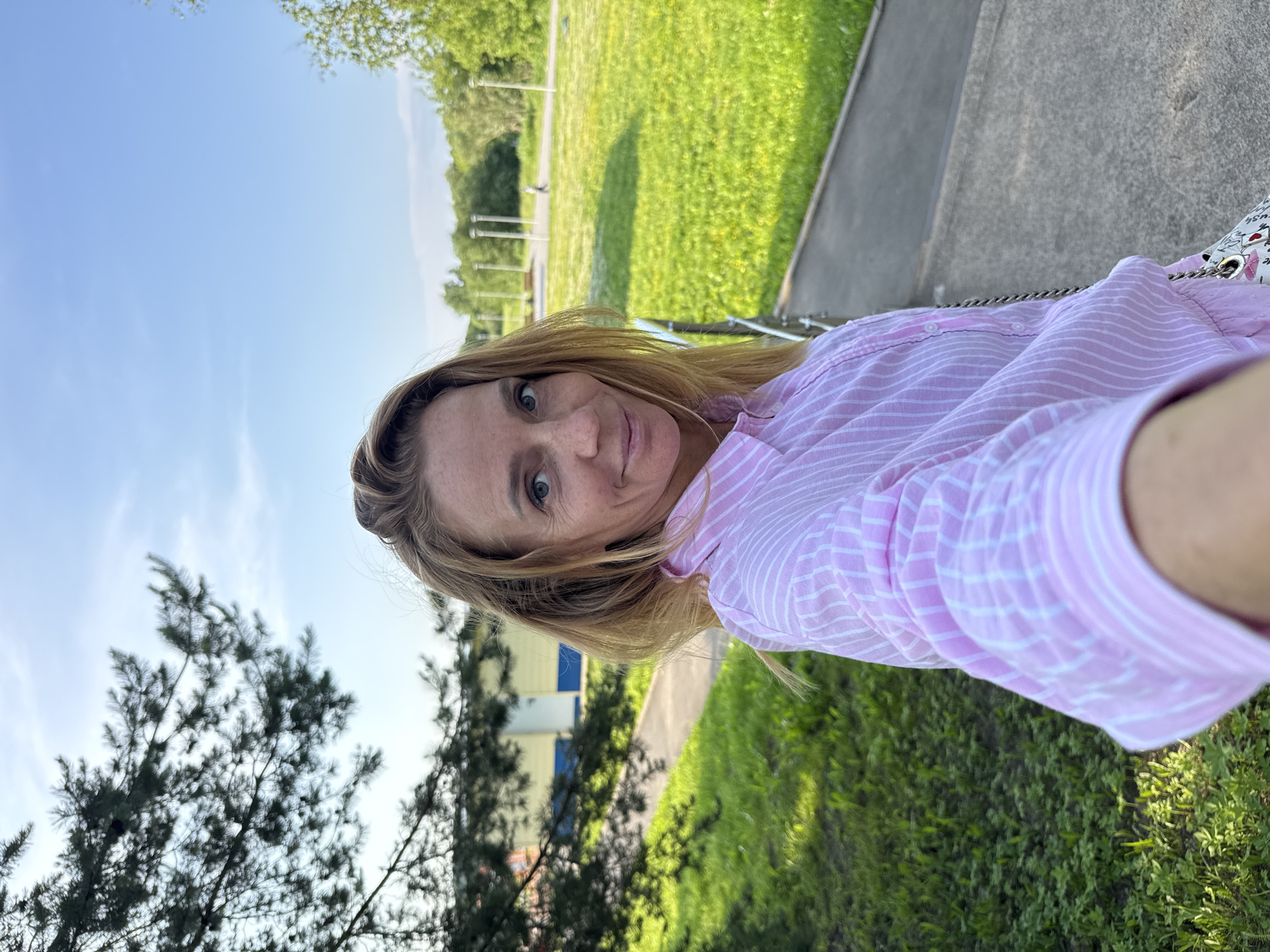 An outdoor, medium shot captured in Kirishi, Russia, shows a woman with blonde hair, wearing a pink and white striped button-up shirt. She is looking directly at the camera with a slight smile. The background is a green grassy field with some trees and a blue and yellow building, possibly a school or sports facility. The lighting suggests it is daytime, with a clear blue sky.
