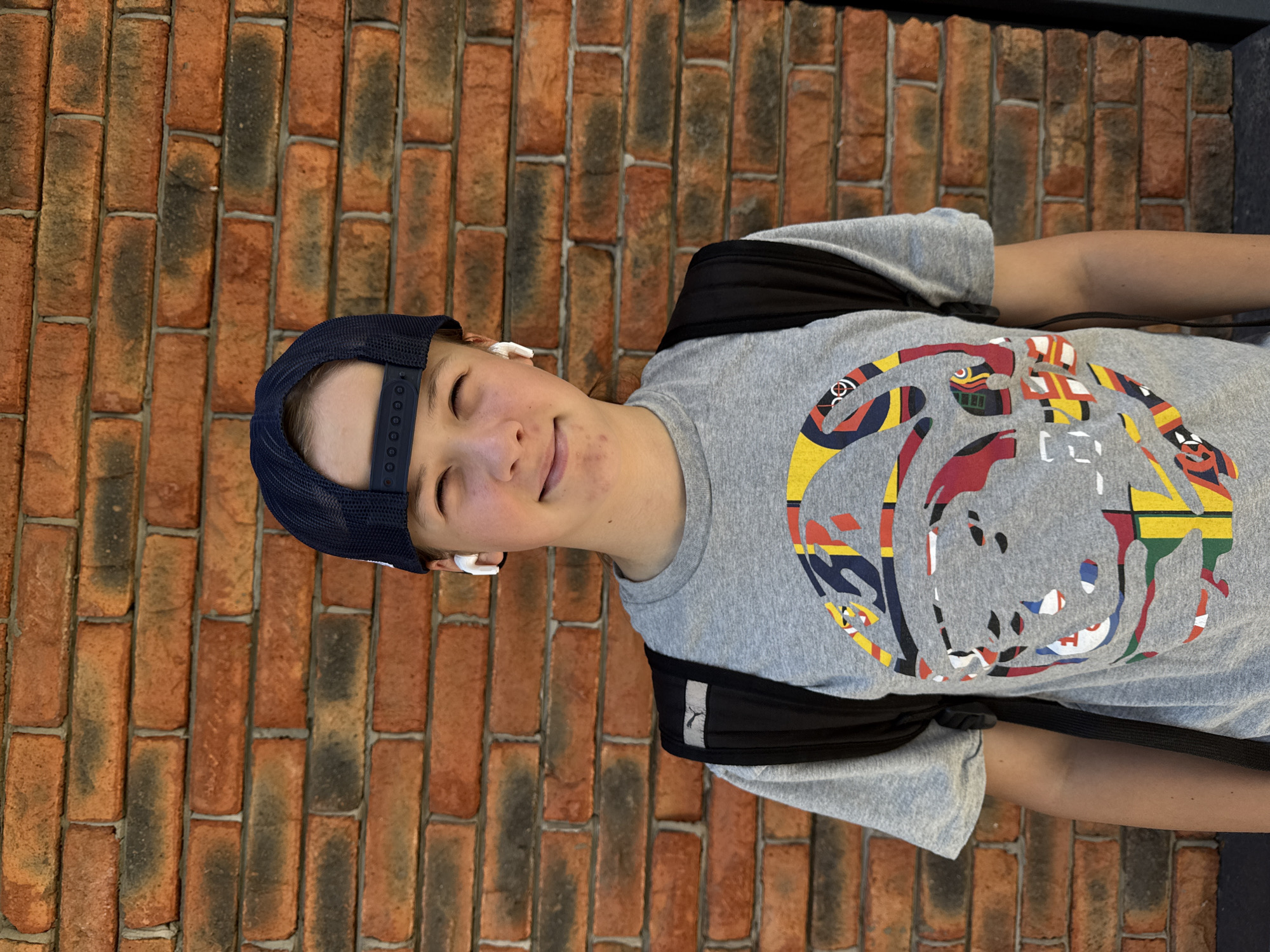 A young person, likely male, stands facing the camera in front of a brick wall. The person is wearing a navy blue baseball cap, a gray t-shirt with a colorful abstract design on the front, and a black backpack. White wireless earbuds are visible in their ears. The person has short, light brown hair and is smiling slightly. The brick wall behind them is made of orange and dark red bricks, laid in a horizontal pattern. The lighting suggests an outdoor setting, possibly during the daytime. The background shows no discernible landmarks to place the image in Chaikovsky, Russia.