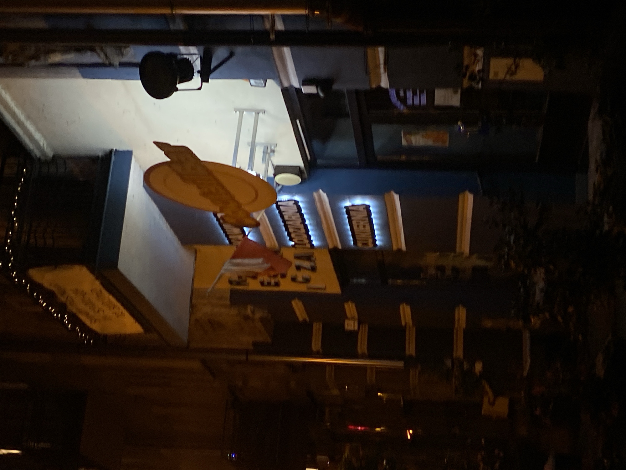 The image shows a storefront at night in Nowy Sącz, Poland. The storefront has a blue facade with yellow and white trim. There are signs with illuminated text on the building. One sign reads "CUKIERNIA", which means "confectionery" in Polish. Another sign, partially visible, appears to read "LODZIARNIA", meaning "ice cream shop". A Polish flag is also visible, hanging from the building. There is a round, yellow decorative element above the entrance. A black floodlight is mounted on the wall to the right of the entrance. The scene is dimly lit, suggesting it is nighttime. The ground in front of the store is covered with foliage, and there are Christmas lights strung above the awning of the building. The overall impression is of a closed business at night.