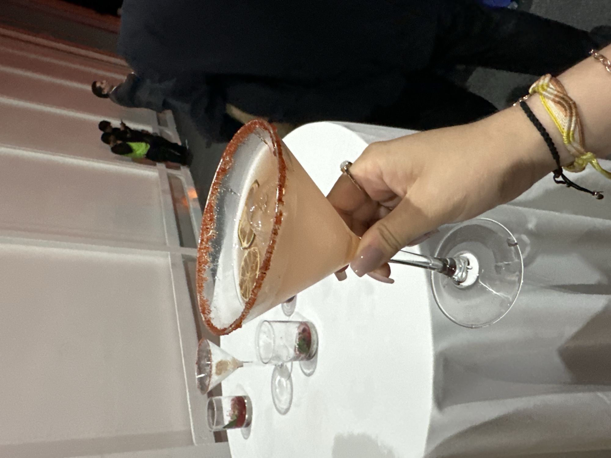 A person holds a martini glass with a reddish rim in their right hand, with two bracelets visible on their wrist. The glass contains a light-colored liquid and what appears to be slices of fruit. On the white table, two other stemmed glasses and two short glasses containing red berries are positioned. In the background, blurred figures of people are visible. This scene could plausibly be taking place at a bar in Mexico City, Mexico.