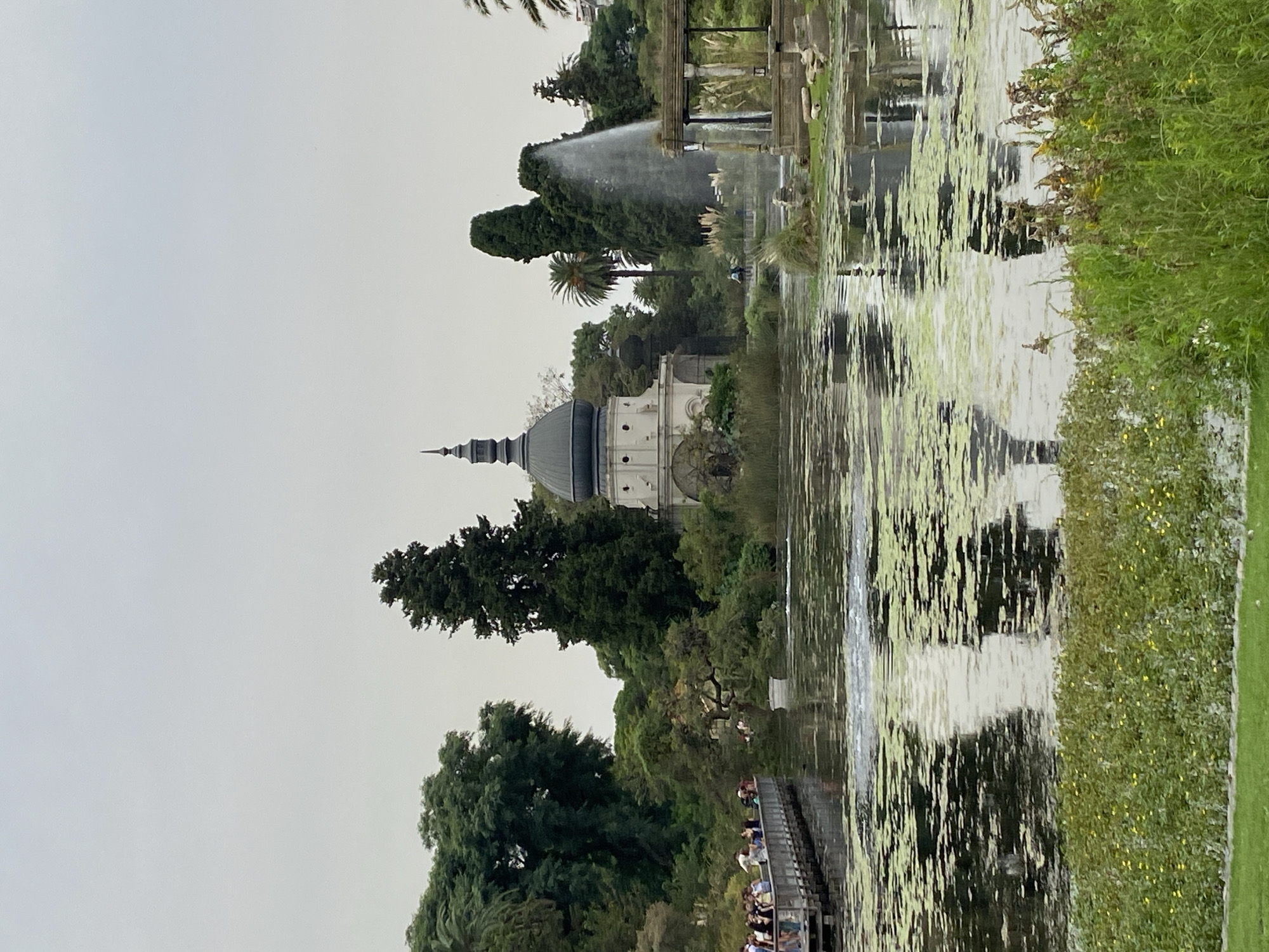 The image shows El Rosedal park, located in the Palermo neighborhood of Buenos Aires, Argentina. In the foreground is a body of water with algae and reflections. On a wooden pier near the water, a group of people is gathered. In the background, a building with a dark dome is visible amidst trees and foliage. A fountain is also seen spraying water. The sky is overcast.