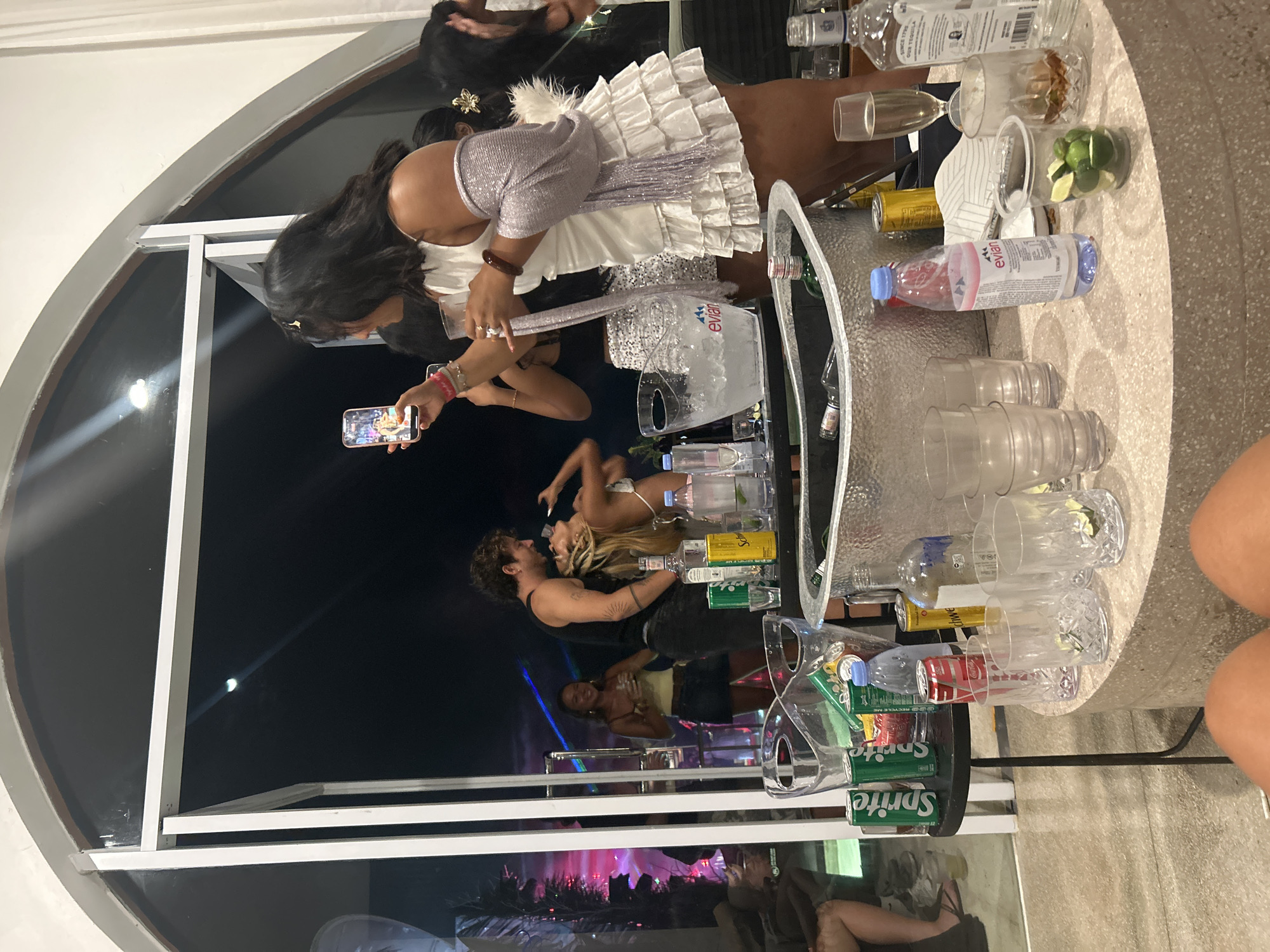 The image captures a lively party scene indoors, possibly at a beach club or a private villa in North Kuta, Indonesia. Several individuals are present, engaged in various activities. In the foreground, a woman in a white ruffled dress is holding a glass and looking towards a person on her right. To her left, another woman is holding a smartphone, taking a picture. Behind them, through a large arched window or doorway, a couple is visible dancing, with colorful lights suggesting a party atmosphere outside. On a circular counter in the foreground, an assortment of drinks and glasses are laid out, including bottles of spirits, cans of soda, and a bucket filled with ice. The overall ambiance is festive and energetic, with people socializing and enjoying themselves.