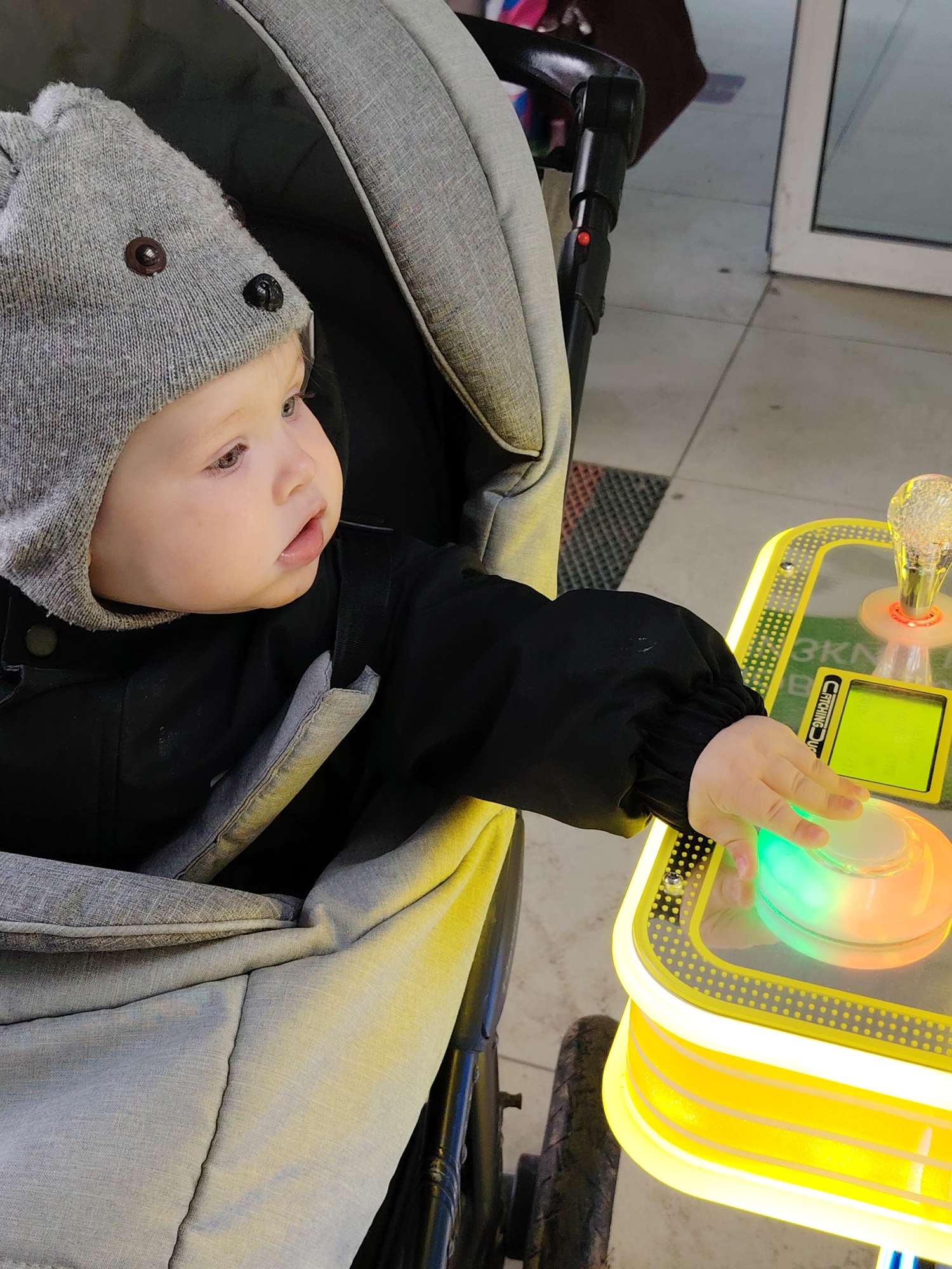 A baby wearing a grey knit hat with button eyes is seated in a stroller, interacting with an illuminated arcade game. The game is yellow with a clear plastic top, featuring a large, circular button that emits a green and orange glow. A small digital screen is visible on the game's surface. The stroller is grey and black. The background suggests an indoor setting with light-colored flooring and a glass door. The location is Chaikovsky, Russia.