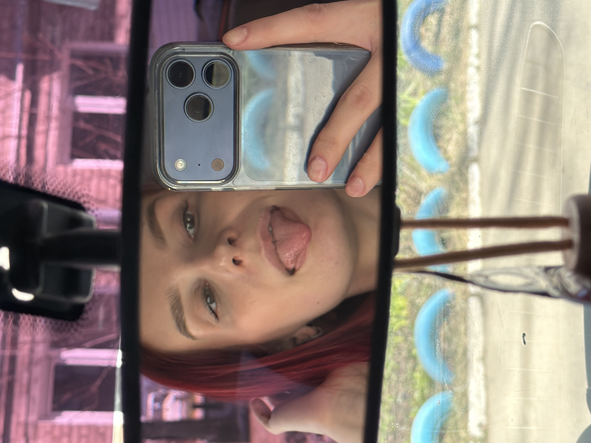 The image depicts a selfie taken in the rearview mirror of a vehicle. A young woman with red hair is visible in the mirror's reflection. She is holding a smartphone with multiple camera lenses. Outside the car, a blurry scene includes buildings, possibly in Chaikovsky, Russia, given the user's request. The exterior view through the car window shows a blue semi-circular design feature. The overall scene suggests a casual snapshot taken inside a car.