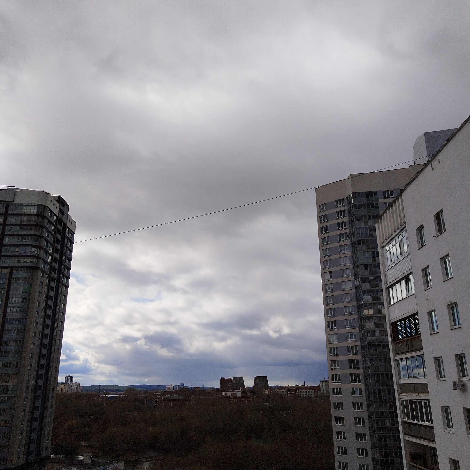 The image shows a cityscape under an overcast sky in Yekaterinburg, Russia. In the foreground, two tall apartment buildings are visible on the left and right sides of the frame. The building on the left has a dark facade with numerous windows. The building on the right is white with visible balconies and windows. A thin wire is stretched between the two buildings.
In the background, beyond the buildings, there is a landscape of trees and lower buildings. Some buildings with industrial design can be seen in the distance, and slightly further, a hint of hills or mountains is visible. The sky is filled with gray and white clouds, indicative of possible inclement weather.