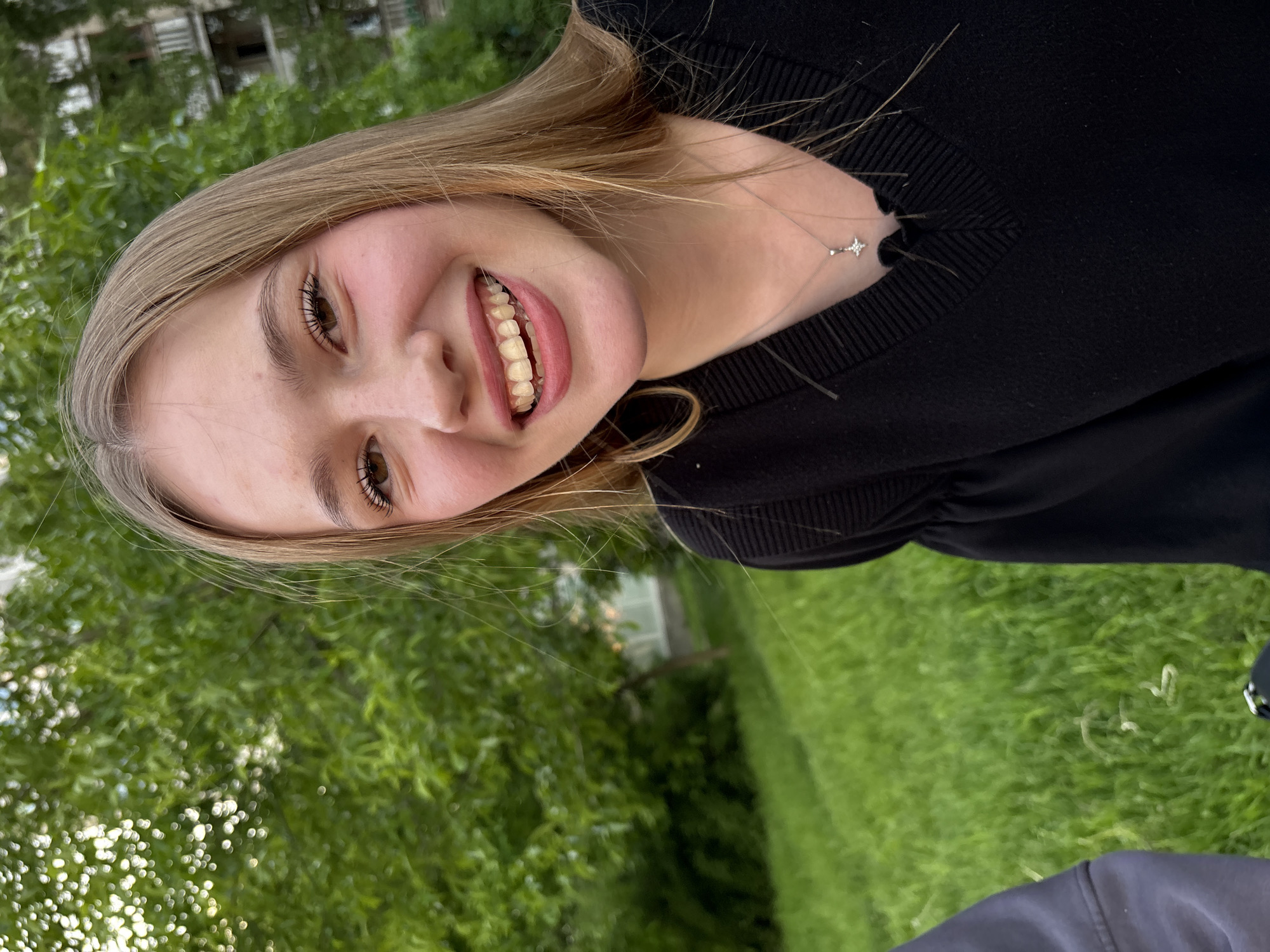 The image captures a young woman smiling widely and looking directly at the viewer. She has light brown hair parted on the side and brown eyes. Her smile reveals an orthodontic appliance on her teeth. She is wearing a black, ribbed V-neck sweater. A delicate silver chain with a small, sparkling star-shaped pendant is visible around her neck, resting on her collarbone.
The setting is outdoors, dominated by a background of vibrant green grass and trees, suggesting a park or garden. In the distance, through the foliage, faint structures resembling buildings can be seen, hinting at an urban or suburban environment in Saint Petersburg, Russia. The lighting appears soft, indicating it is daytime. The woman's expression is cheerful and happy. There is no visible text in the image.