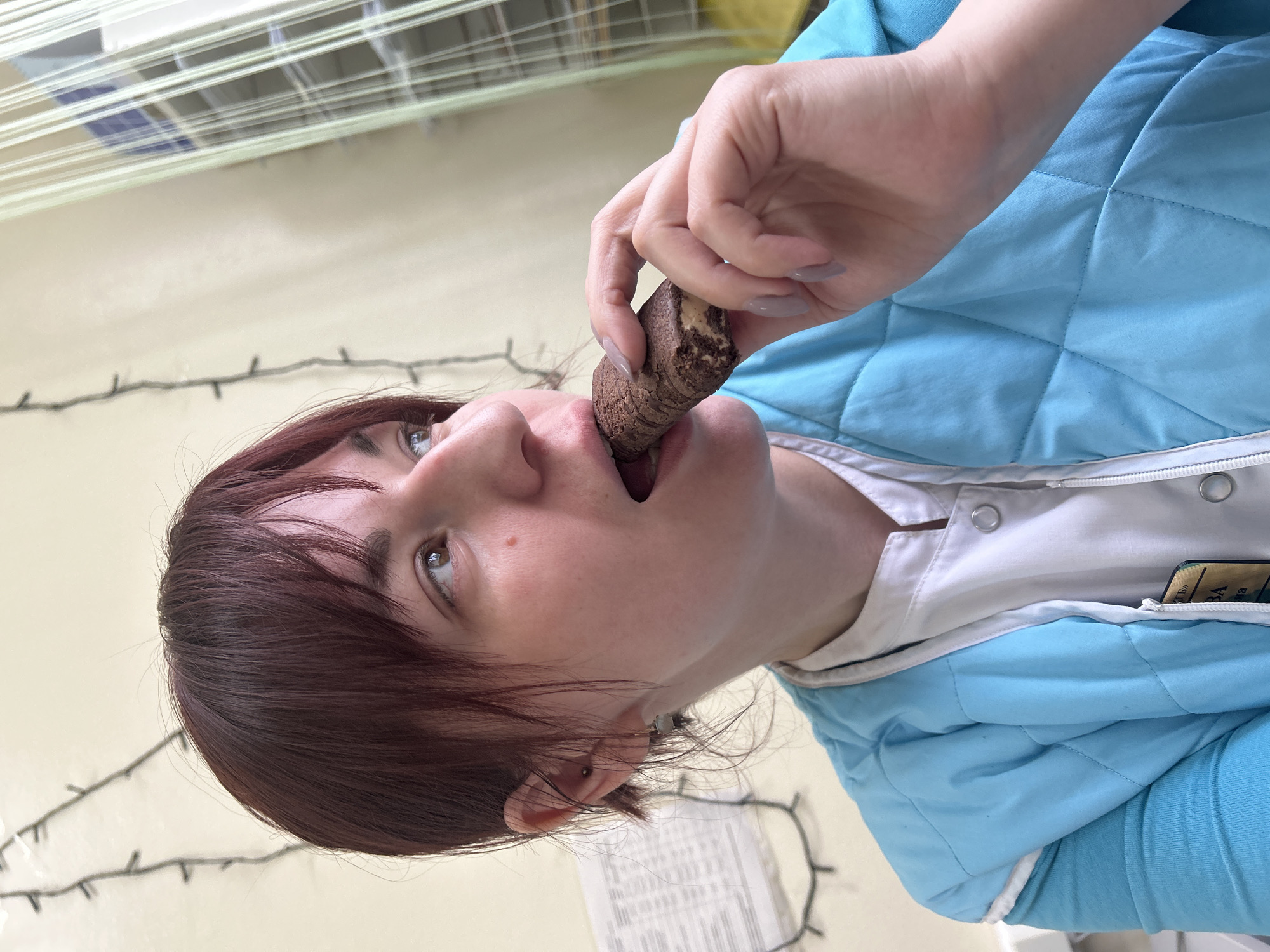 An indoor, close-up shot captures a person with reddish-brown hair eating a snack bar. The person, viewed from a low angle, has their mouth open with the dark brown, possibly chocolate-flavored, bar partially inserted. Their right hand, adorned with light-colored nail polish, holds the bar. Their eyes are cast upwards.
The person wears a light blue quilted jacket over a white garment, possibly a uniform shirt, which features visible snap buttons. Tucked into a pocket of the white garment, a dark card with some yellow text, partially visible as "ВА", can be seen. A small, simple stud earring is visible in the person's left ear.
In the background, a light-colored wall is visible, with a string of small, dark lights running horizontally across it. Above the person's head, what appear to be horizontal blinds or a window covering are visible against a lighter background. The overall setting appears casual yet functional, possibly a break room or a similar indoor space.