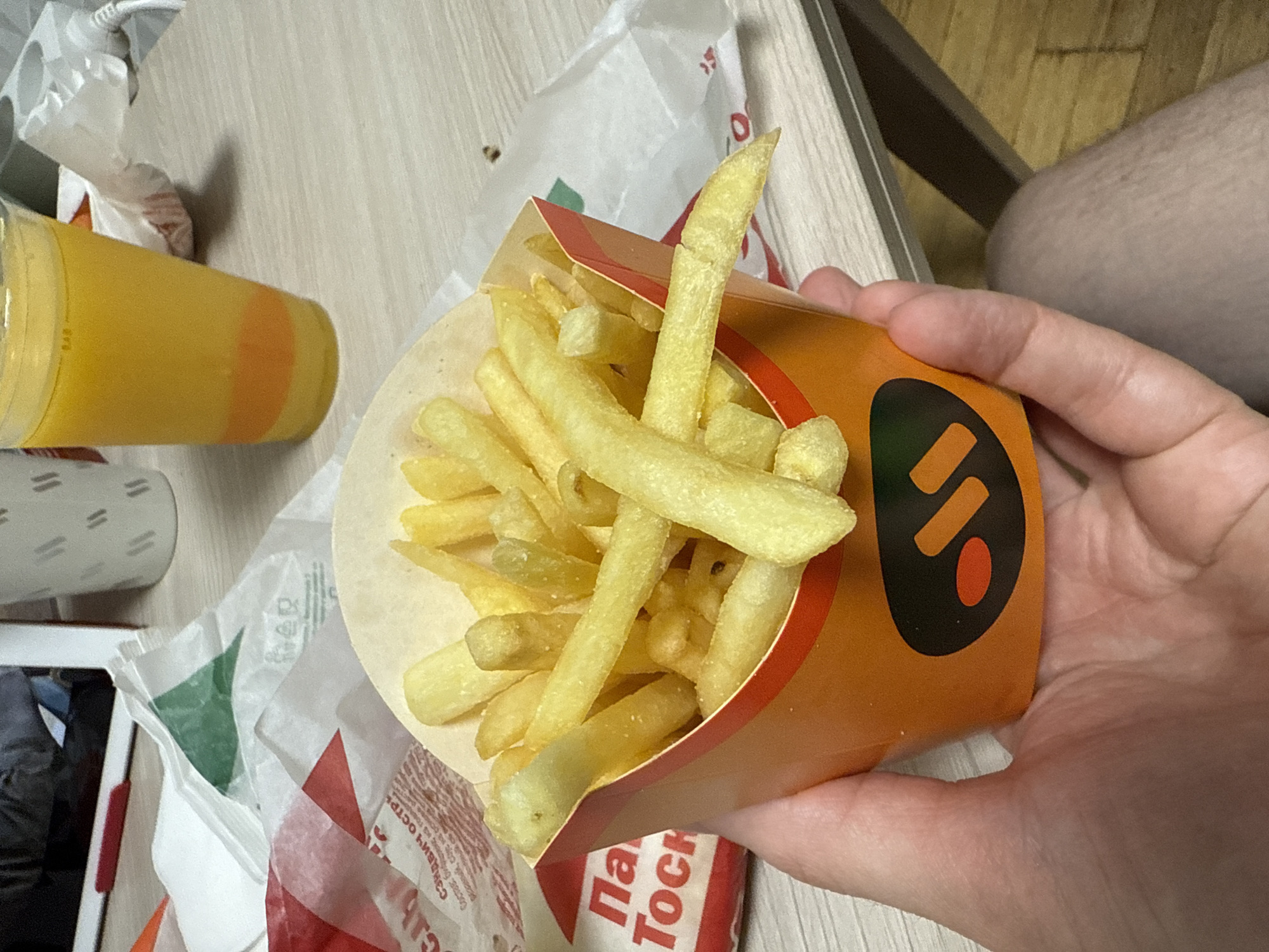 In this overhead shot, a person's hand holds a carton of french fries. The carton is bright orange with a black and red logo featuring a stylized "S" or "5". The fries are golden brown and appear to be freshly cooked. To the left of the carton is a clear plastic cup filled with a yellow liquid, likely juice or a similar beverage. The cup has an orange band around its middle. The items are arranged on a light-colored, textured surface, possibly a table, with some crumpled white paper packaging visible in the background. The lighting is bright, suggesting it's daytime. The overall scene conveys a casual, possibly fast-food dining experience. Visible text on the packaging is partially obscured but includes Cyrillic letters.
