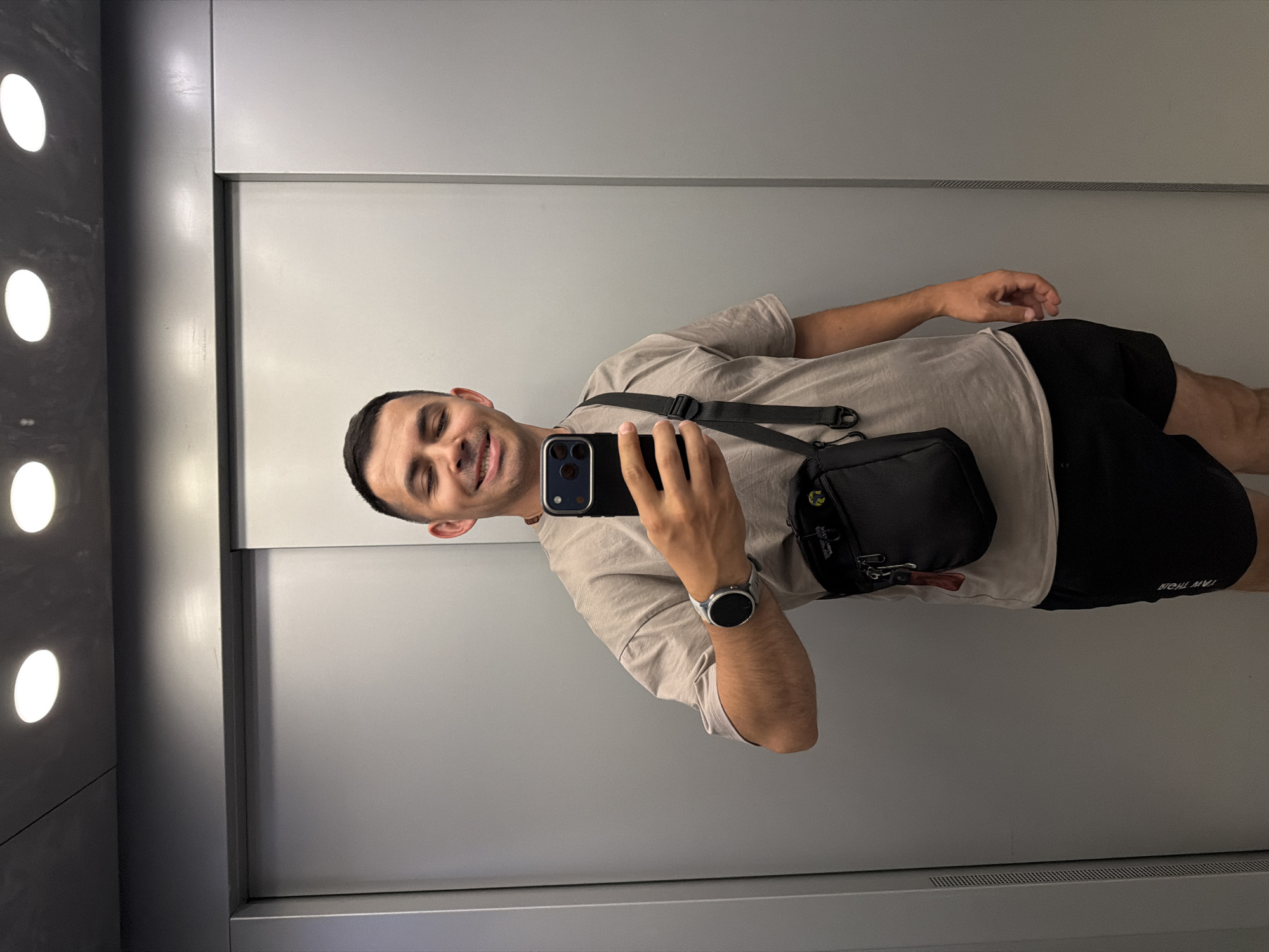 The image shows a man standing in an elevator, taking a selfie. He is wearing a light brown t-shirt, black shorts, a black crossbody bag, and a smartwatch. The elevator has gray walls and two circular lights on the ceiling. The man is smiling with his eyes closed. Based on the provided information, the location is an elevator in the city of Izhevsk, Russia.