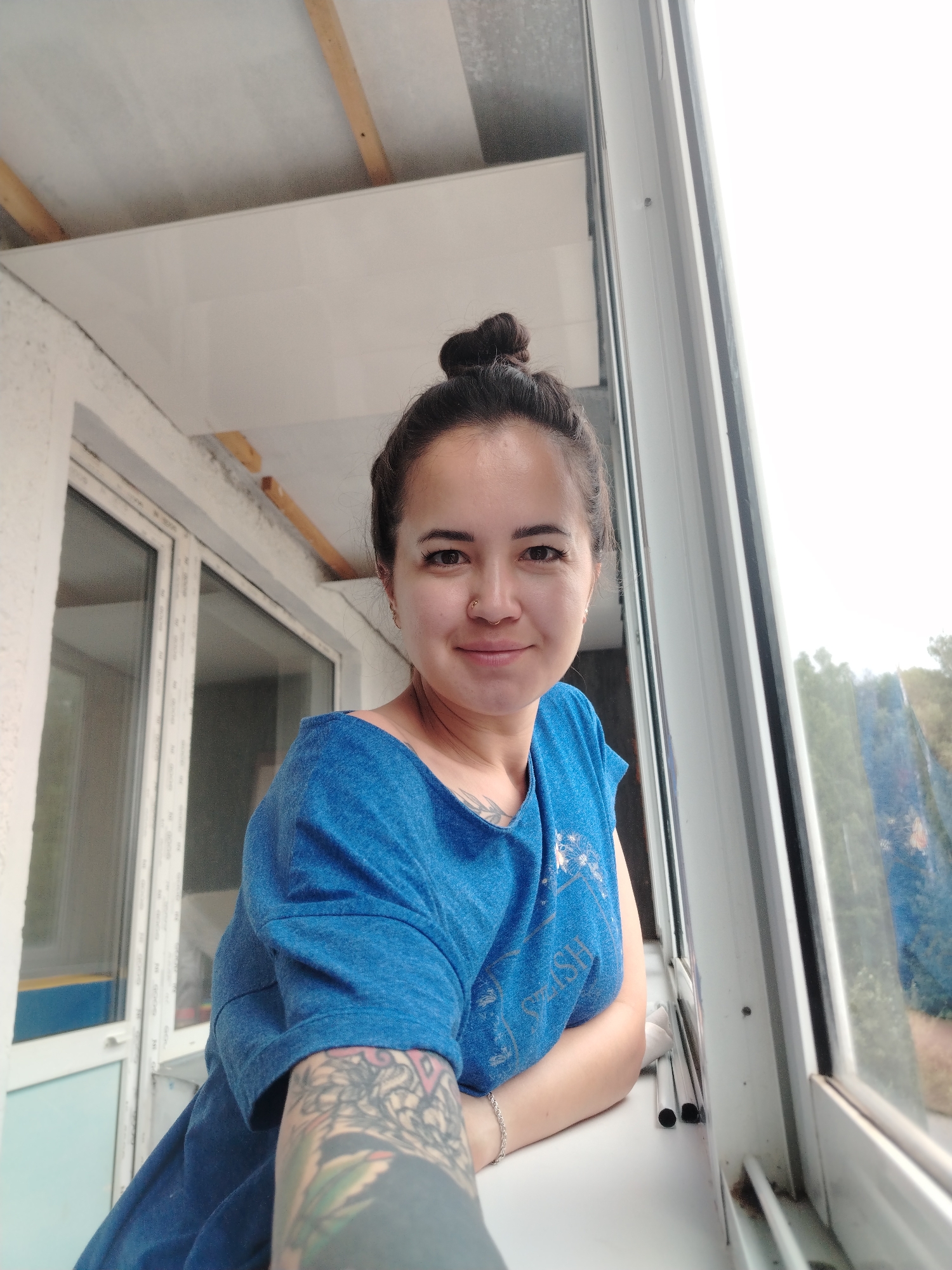 A young woman with dark hair pulled into a bun and a nose ring is sitting on a windowsill. She is wearing a blue t-shirt and has a tattoo on her arm. She is smiling at the camera. The background appears to be a balcony with a window looking out onto a green landscape. The image is taken from a slightly low angle. The scene is likely in a residential area, possibly an apartment building, given the balcony and window setup. The presence of what appears to be a balcony and the overall context suggest a dwelling in a populated area. The mention of Noviy, Russia, places the location specifically.
