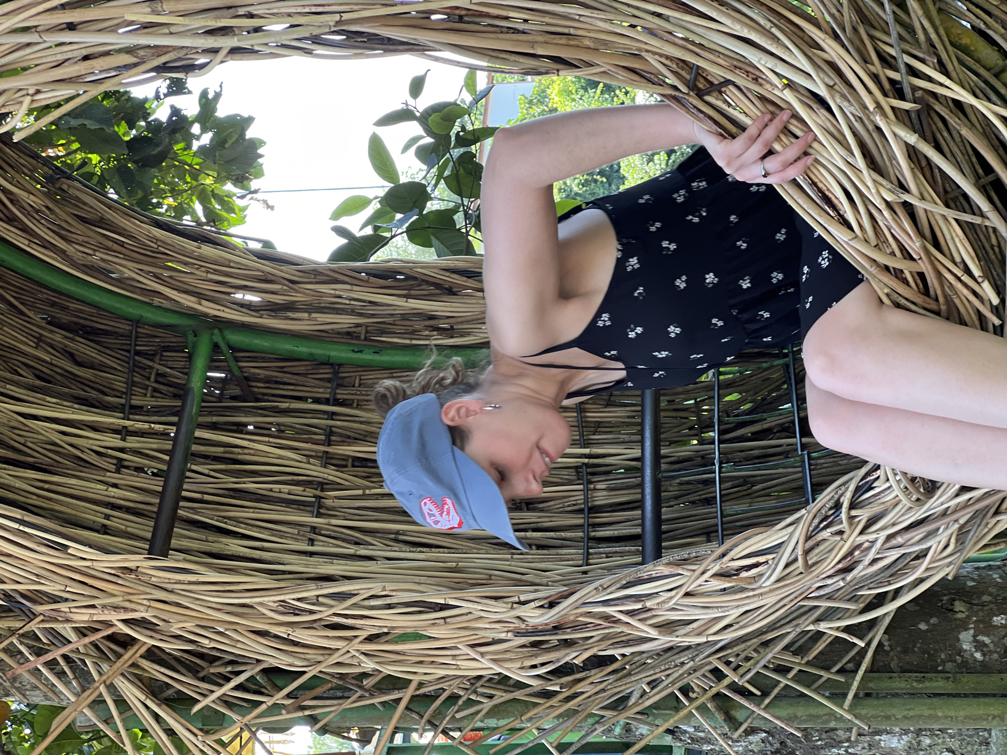 A woman is seated in a large, woven nest-like structure made of natural materials, possibly at a tourist attraction in Sukawati, Indonesia. She is wearing a blue baseball cap with a red and white dinosaur logo, a black dress with a floral pattern, and has a ring on her finger. The nest is situated outdoors, with lush green foliage visible in the background, suggesting a tropical environment. Sunlight filters through the leaves, illuminating the scene. The structure appears to be constructed from various branches and twigs, woven together to form a rounded, enclosed space.