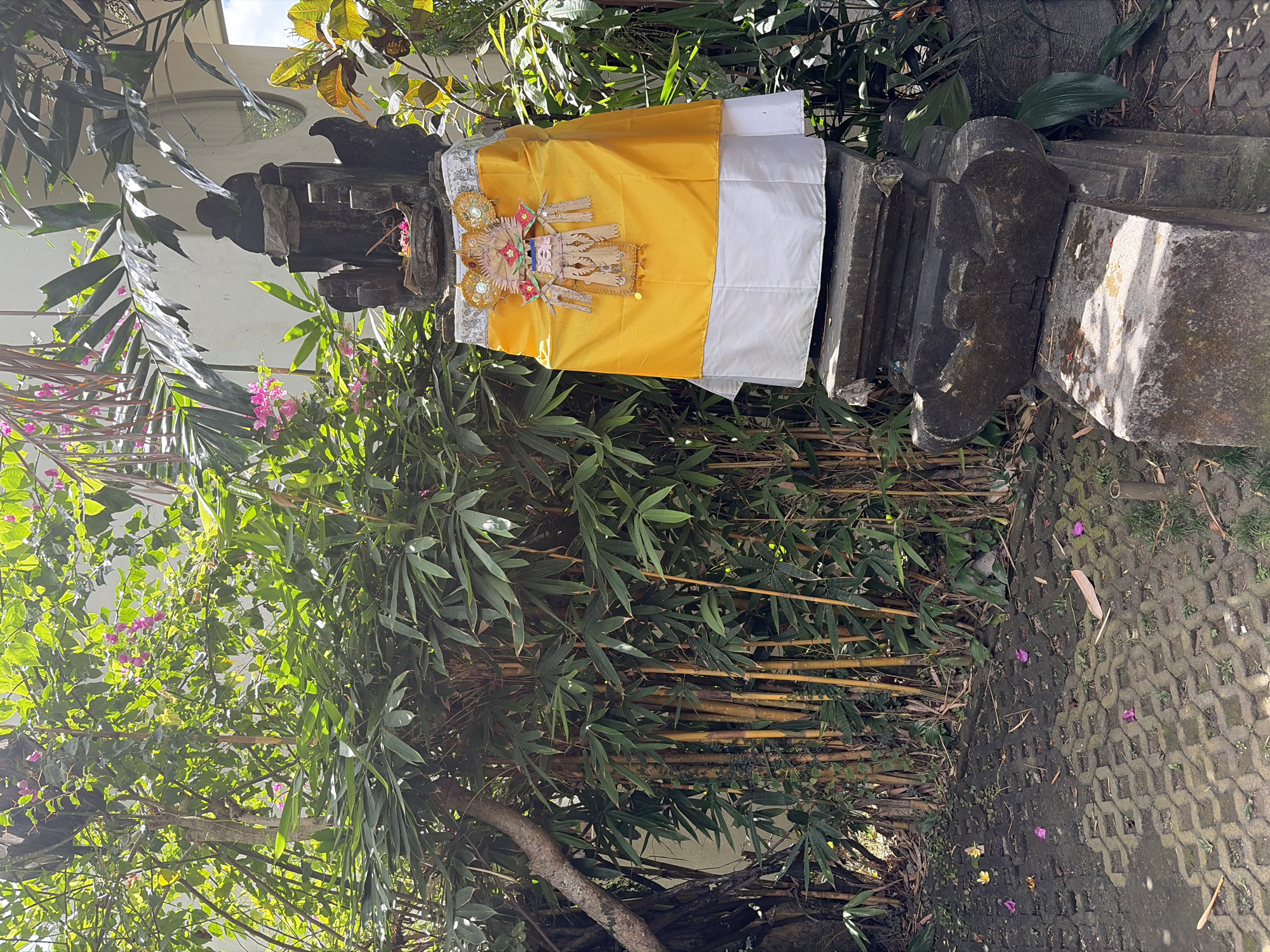 In Mengwi, Indonesia, a stone shrine stands amidst lush greenery. The shrine, intricately carved from dark stone, is adorned with a vibrant yellow cloth, a white cloth, and a decorative offering at its apex. The surrounding environment is rich with plant life, including bamboo and other tropical foliage, some of which bear small pink flowers. The ground in front of the shrine is paved with interlocking bricks, scattered with fallen leaves and a few scattered purple and yellow flowers. Sunlight dapples the scene, highlighting the textures of the stone and the vibrant colors of the fabrics.