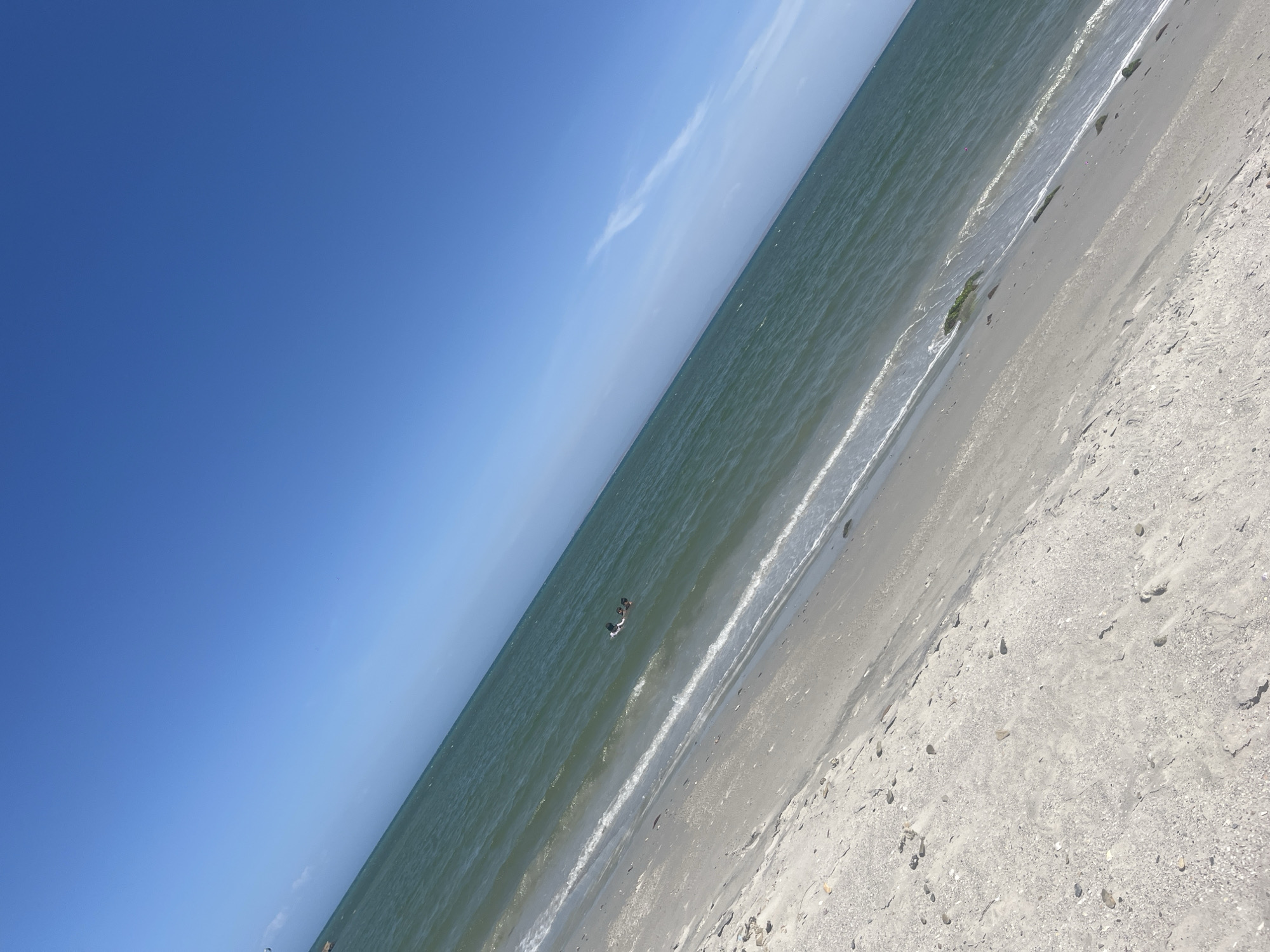 The image shows a beach scene, likely in El Yaque, Venezuela, under a clear blue sky with a few wispy clouds. The water is a muted green color, with small waves approaching the sandy shore. The beach is light gray and appears to be made of fine sand and shell fragments. In the water, there are two individuals. The beach shows scattered footprints.