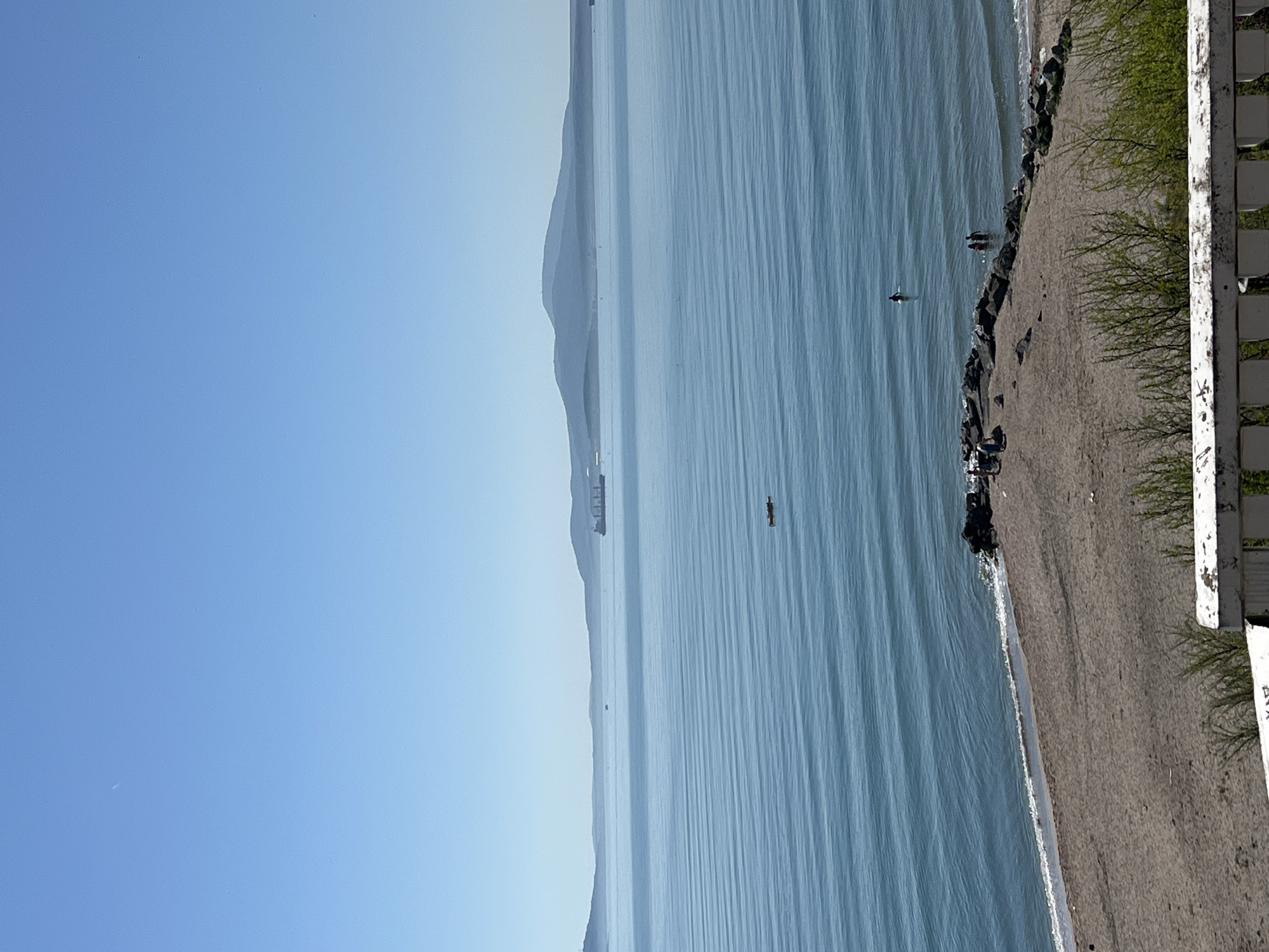 The image captures a coastal scene in Burgas, Bulgaria, featuring a body of water (likely the Black Sea) and a beach. The sky is clear and light blue. The water has a light blue hue with visible ripples and some boats. A few people are seen in the water near the beach. The beach is sandy with some rocky areas along the water's edge. Vegetation, including grass and trees, borders the beach. A weathered white fence appears in the foreground. A large mountain is visible on the left.