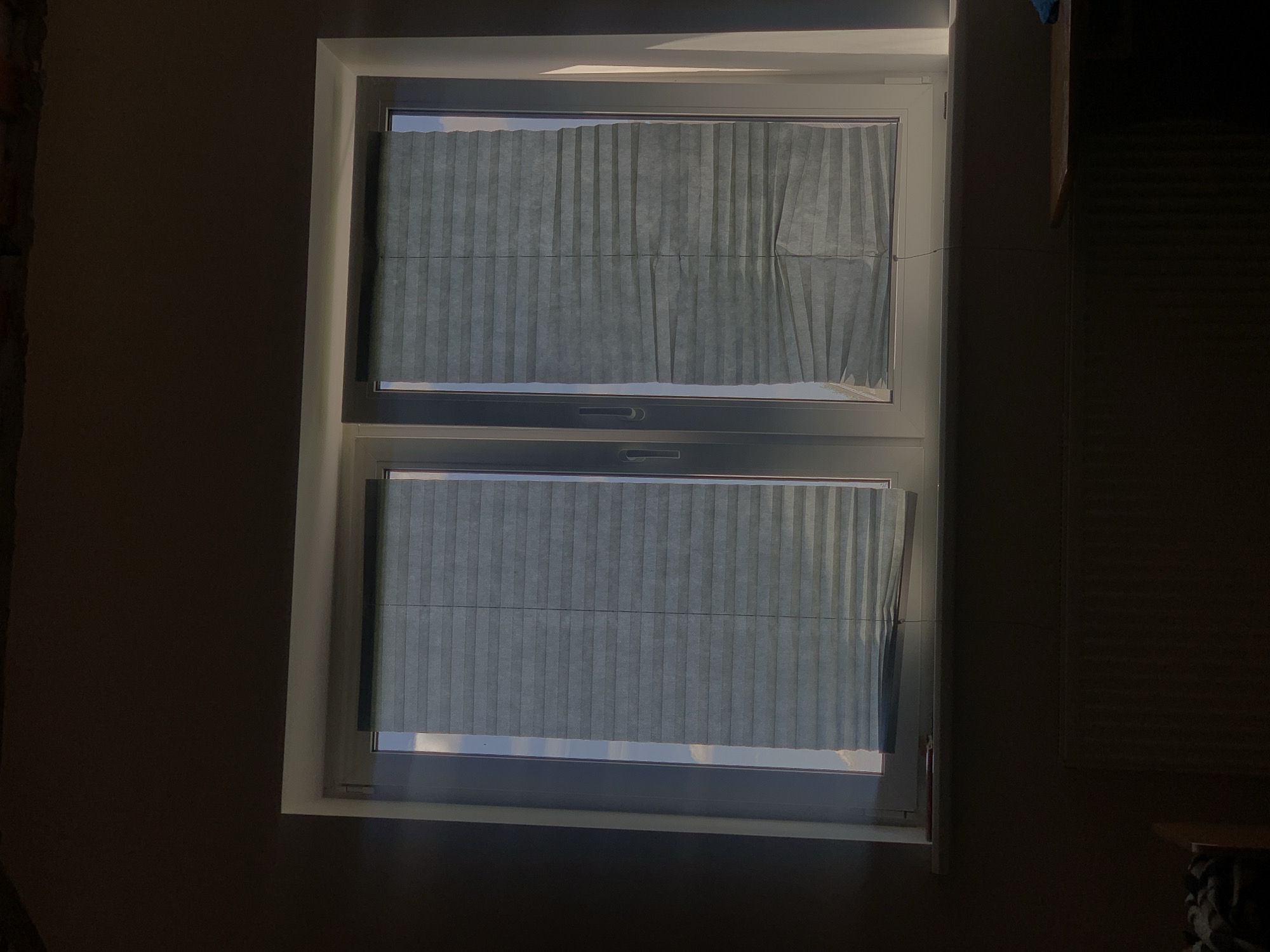 The image depicts a dimly lit room interior, focused on a double-paned window. The window has grey pleated blinds partially drawn, obscuring the view of the sky outside. The left pane has its blinds mostly lowered, while the right pane's blinds are drawn higher, revealing more of the sky. The window is set into a white frame and sits on a windowsill. Below the windowsill, a section of a radiator is visible. The room's walls are dark and appear to be painted or plastered. Sunlight casts a strong shadow across the right side of the window frame and the wall above it, suggesting the sun is low in the sky. The overall impression is of a domestic space, likely a residential apartment. The location in Minsk, Belarus cannot be determined from the visual information provided.