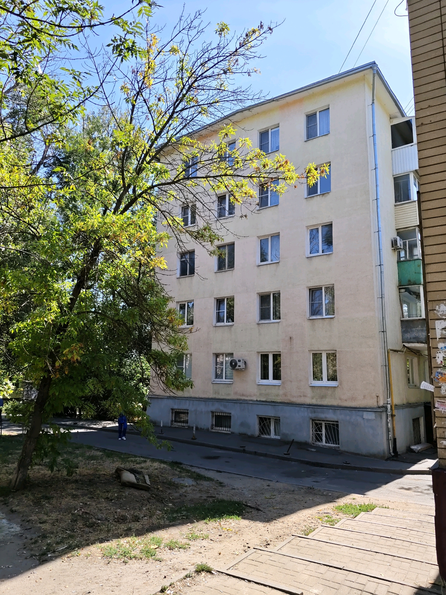 A light beige apartment building is visible from the side, with a tree in the foreground casting a shadow. The building has at least five floors visible, with numerous windows and balconies. The ground level of the building is painted gray and has barred windows. There is an air conditioning unit mounted on the wall of the building. In the middle ground, a person wearing a blue top and blue pants is walking on a paved path. The ground in the foreground is dirt and grass. The scene appears to be in an urban environment, possibly a residential area.