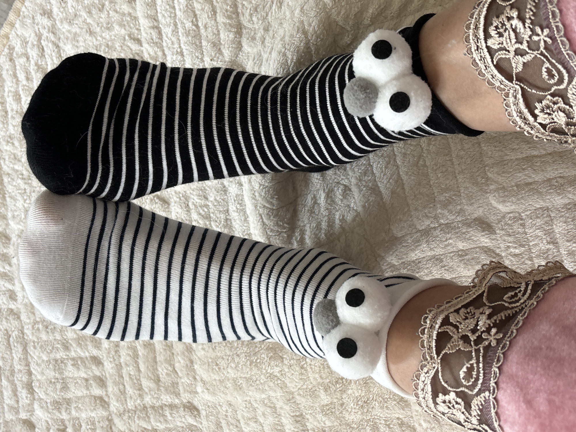 A downward shot shows two legs wearing striped socks with googly eyes. The top sock is black with thin white stripes and a black toe. The bottom sock is white with thin black stripes and a white toe. Both socks have two large white googly eyes and a smaller grey felt nose. The legs are resting on a textured white fabric surface. The leg on the right has part of a pink garment visible, trimmed with a beige lace detail. There are no indications that the scene is in Chaikovsky, Russia.