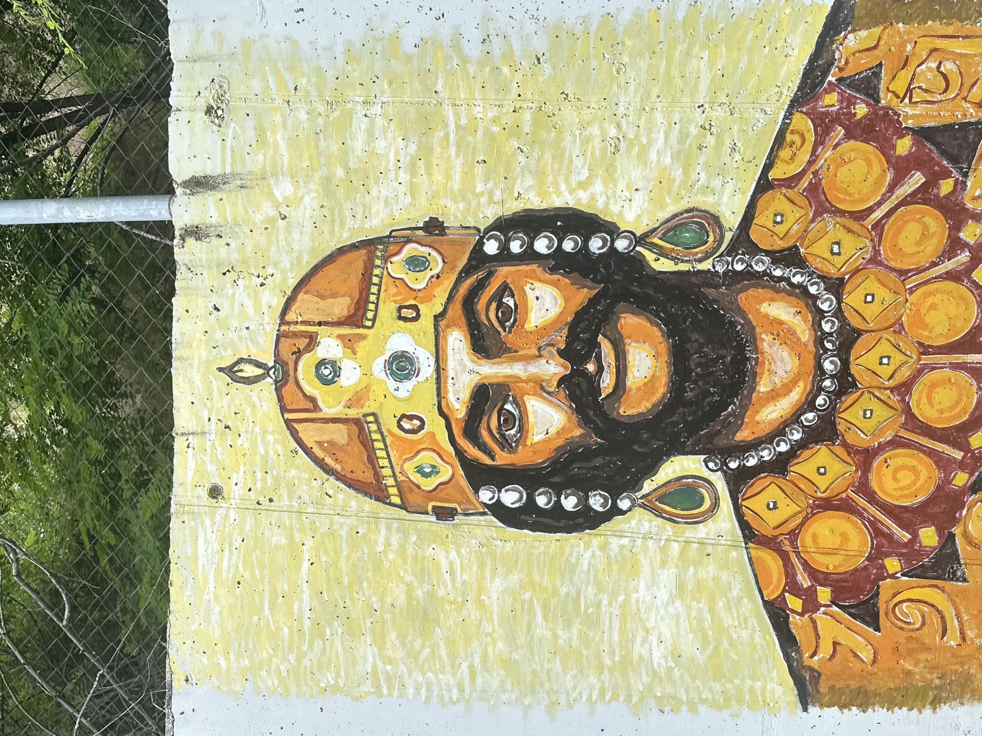 An outdoor mural depicts a stylized portrait of a regal figure, possibly a king or emperor, painted on a concrete wall. The figure, facing forward, has dark skin, a prominent dark beard and mustache, and large, dark eyes. They wear an elaborate golden and orange crown or headpiece adorned with white flower-like patterns and small white circles resembling pearls or gems. A necklace of white pearls drapes around the neck, accompanied by green teardrop-shaped earrings. The attire is richly patterned with intricate designs in shades of gold, orange, and reddish-brown, creating a sense of grandeur.
The artwork is set against a textured yellow background, showing visible brushstrokes and some signs of wear on the concrete surface, including small dark specks and horizontal lines that might be joints or damage. To the left of the mural, a chain-link fence is visible, along with a grey metal pole and lush green foliage, indicating an outdoor, possibly urban or semi-urban, environment. The lighting suggests it is daytime. There is no discernible text within the artwork or its immediate surroundings. This mural is located in Asenovgrad, Bulgaria.