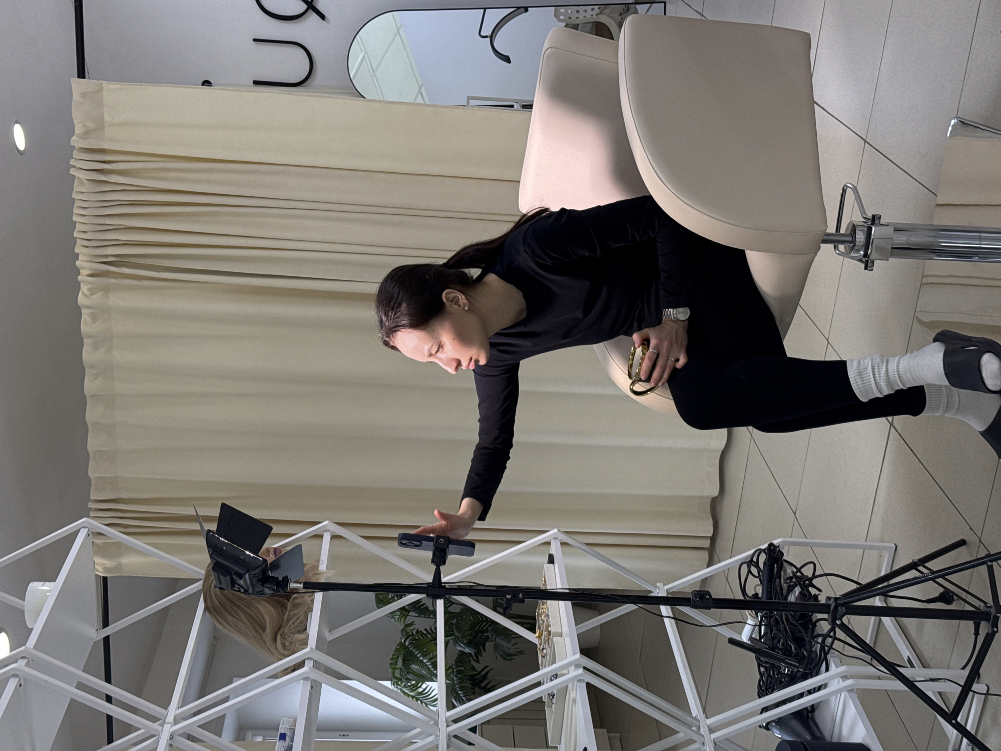 An indoor scene, likely a studio or salon in Chaikovsky, Russia, shows a woman setting up equipment for what appears to be a recording or photoshoot. The image is captured from a slightly elevated perspective.
The primary subject is a woman with dark hair, wearing a black long-sleeved top, black leggings, white socks, and black sandals or slides. She has a watch on her left wrist and rings on her fingers, holding a gold-colored object in her left hand. She is leaning forward from a light beige salon chair, extending her right arm towards a smartphone mounted on a tripod. Her expression is focused, indicating concentration on the task.
The immediate setting includes a professional styling chair. In front of her, a black tripod holds a smartphone in a mount, positioned next to a white, geometric shelving unit. On the shelving unit, a blonde wig head is visible, and further down, possibly some products or jewelry. A light source, perhaps a small LED panel with barn doors, is also attached to the tripod or a related stand.
To the left, light beige Roman blinds or curtains cover a window or wall, with a recessed light visible on the ceiling above. On the wall above the curtains, a black, irregularly shaped mirror and some large, black curved elements (possibly part of a logo or signage) are discernible. The floor is tiled in a light, neutral color. Near the base of the tripod, a jumble of black cables and wires suggests active equipment. The overall ambiance is clean and professional, typical of a space used for beauty or content creation.