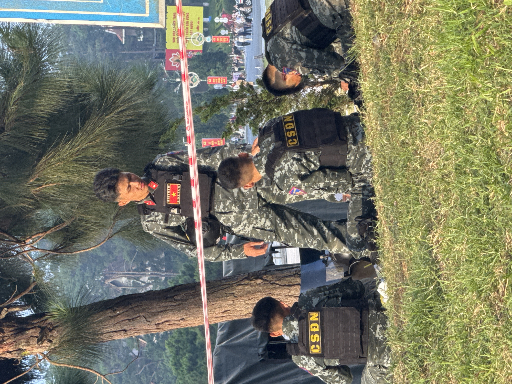 Four individuals in camouflage uniforms with tactical vests are visible. The vests have the text "CSDN" in yellow lettering. One individual is standing and looking down, appearing to give instructions to the others. Three individuals are in a crouching or kneeling position on grassy ground. A red and white striped barrier tape is present in the foreground. In the background, a crowd of people, vehicles, and buildings are observable, suggesting a public urban setting. The presence of trees and foliage indicates an outdoor environment, likely a park or roadside area within the city of Da Lat, Vietnam. The individuals' attire and gear suggest a law enforcement or security operation.