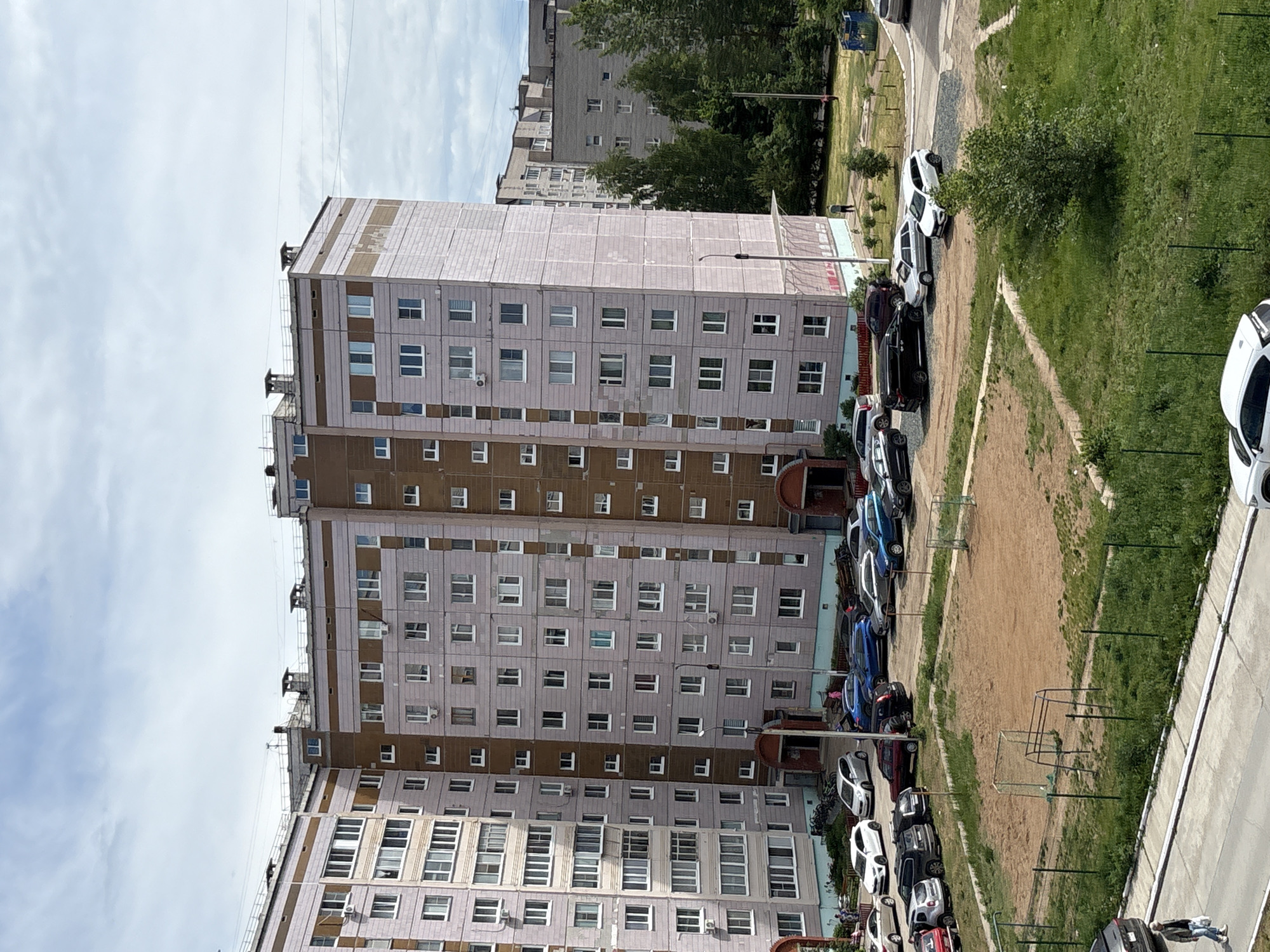 The image shows a residential scene in Chaikovsky, Russia, dominated by a tall, multi-story apartment building with a gray and brown facade. The building has rows of windows, and there are visible structures on the roof. Alongside the building, a row of parked cars of varying colors lines the street. An unpaved field or yard is situated next to the parking area, with what appears to be a rudimentary soccer goal present. Beyond the field, there is a paved walkway and a patch of greenery with a few trees. Another apartment building is visible in the distance, partially obscured by trees. The sky is partly cloudy. There are people on the sidewalk.