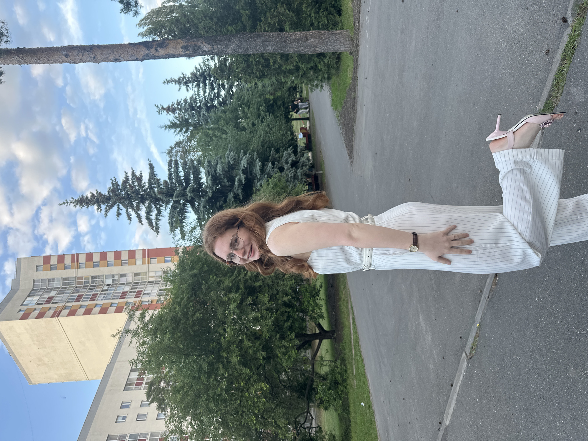 A young woman with long, reddish-brown hair and glasses is posing for a photograph in an outdoor setting. She is wearing a light-colored (likely white or off-white) striped sleeveless jumpsuit or dress with a matching belt and pink high-heeled sandals. A watch is visible on her left wrist, and her right hand is placed on her hip. She is smiling directly at the camera.
The scene features a paved path or road in the foreground and midground, bordered by green grass and numerous trees, including tall pine or evergreen trees and deciduous trees with lush foliage. In the background, a multi-story building with a distinctive facade is visible. The building has a checkered pattern of red, yellow, and white or light-colored panels on its upper floors, with a solid lighter-colored section below. The sky is partly cloudy with patches of blue. The lighting suggests it is daytime, possibly late afternoon or early evening. The overall atmosphere appears pleasant and relaxed. There is no visible text in the image. The location context is Kazan, Russia.