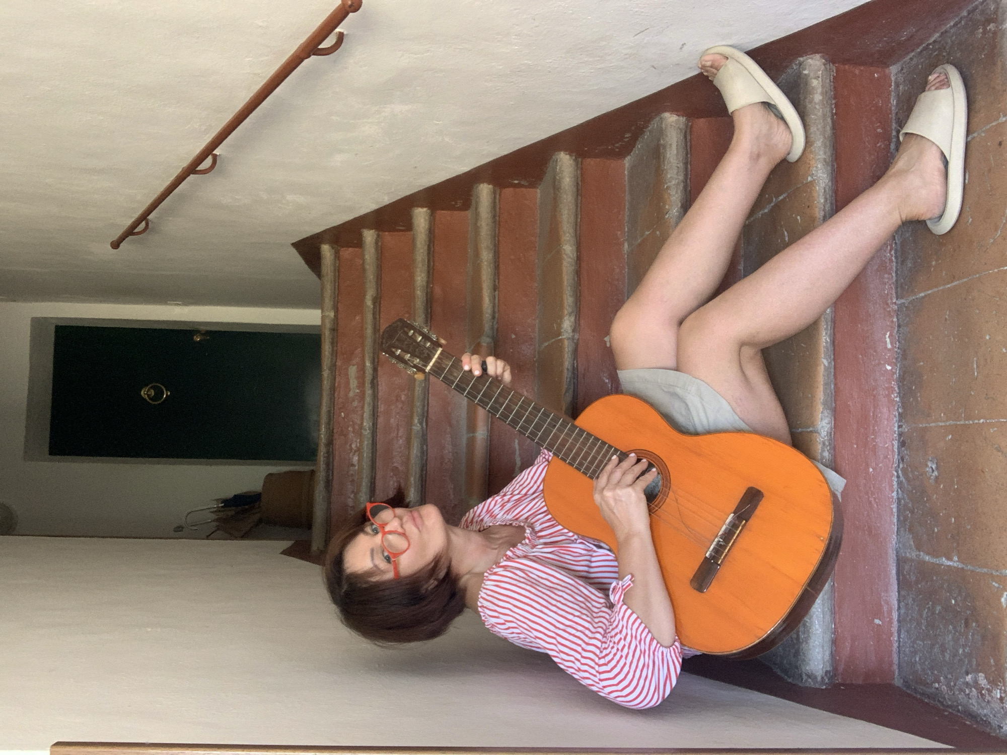 In San Quirico d'Orcia, Italy, a woman sits on a staircase with an acoustic guitar. She is wearing a red and white striped shirt and a light-colored skirt, and bright red-rimmed glasses. The staircase, made of reddish-brown stone and concrete, ascends a narrow passageway. A dark green door with a brass knocker is visible at the top of the stairs, and a wooden handrail runs along the right wall. The woman is holding the guitar on her lap and appears to be playing it, with her fingers positioned on the fretboard. Her gaze is directed towards the camera.
