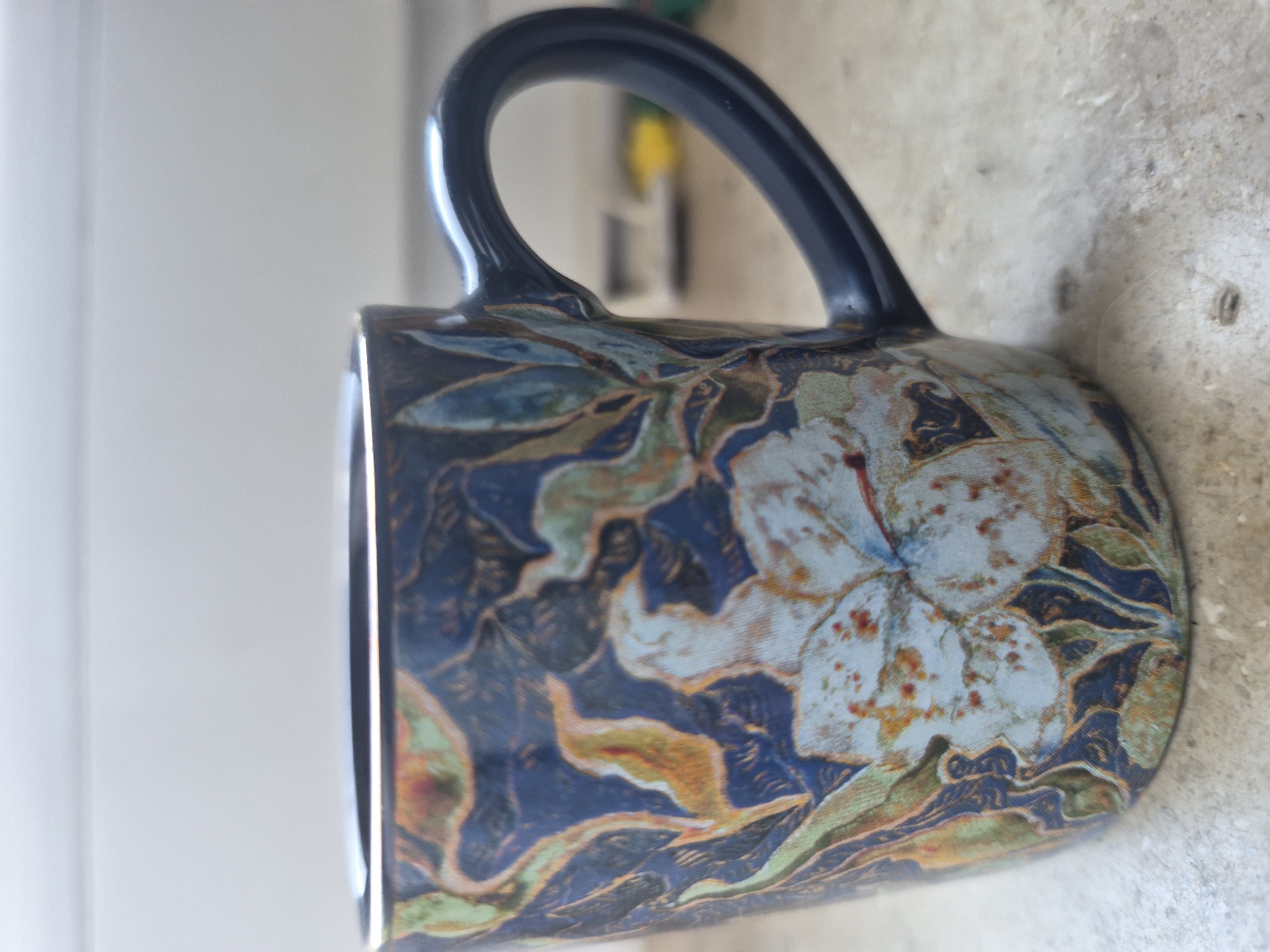 This media file is a close-up, sideways shot of a dark blue mug with a floral pattern. The mug has a dark blue handle and its exterior is decorated with large, light blue flowers with reddish-orange speckles, interspersed with dark blue leaves and foliage in shades of green, yellow, and orange. The floral design has a somewhat painterly or watercolor aesthetic. The mug is resting on a textured surface that appears to be concrete or stone. The background is out of focus, with a bright, possibly window-lit area on the left and a blurred, yellow and green object further back on the right, suggesting an indoor environment. There is no visible text. The lighting appears to be natural daylight, casting soft shadows.