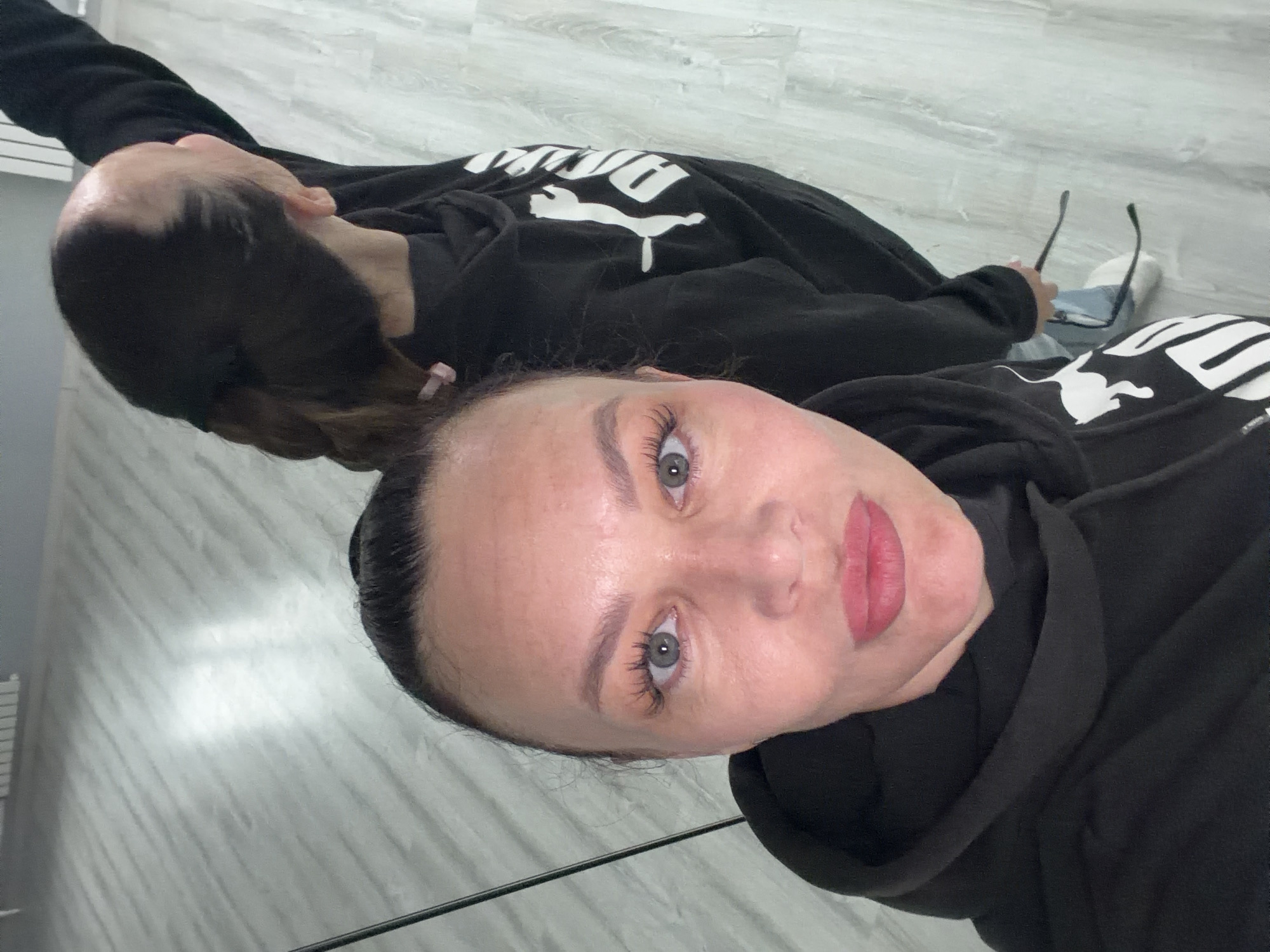 The image shows a selfie of a fair-skinned woman wearing a black Puma hoodie. She has light eyes and pink lipstick. The reflection in the mirror behind her reveals another angle of her and the hoodie, along with a glimpse of blue jeans, white sneakers, and eyeglasses. The background is a light-colored, possibly tiled floor, suggesting an indoor setting. The presence of what looks like a heating register on the left wall may suggest this took place in an apartment or dwelling. While the prompt suggests it takes place in the city of Chaikovsky, Russia, this cannot be confirmed or denied from just the image.