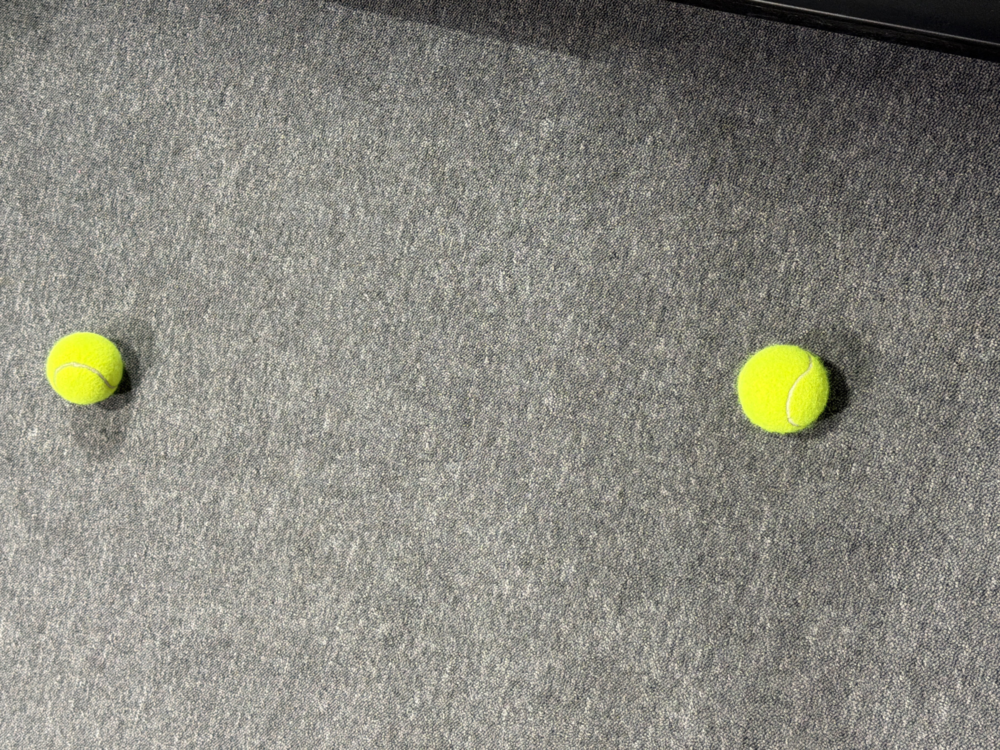 Two tennis balls are positioned on a gray carpet. The balls are located in the left and right areas of the image. There are no people or other objects visible. The location is undetermined.