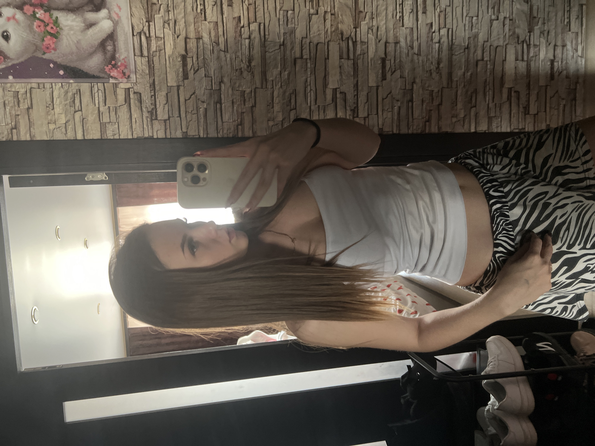 A young woman is taking a selfie in a mirror. She is wearing a white tank top and zebra-striped shorts. Her hair is long and brown. The background includes a brick-patterned wall, a stuffed animal with pink flowers, and a clothing rack with shoes. The location appears to be indoors, possibly in an apartment or dressing room, but the city of Noviy, Russia, cannot be confirmed from this image.