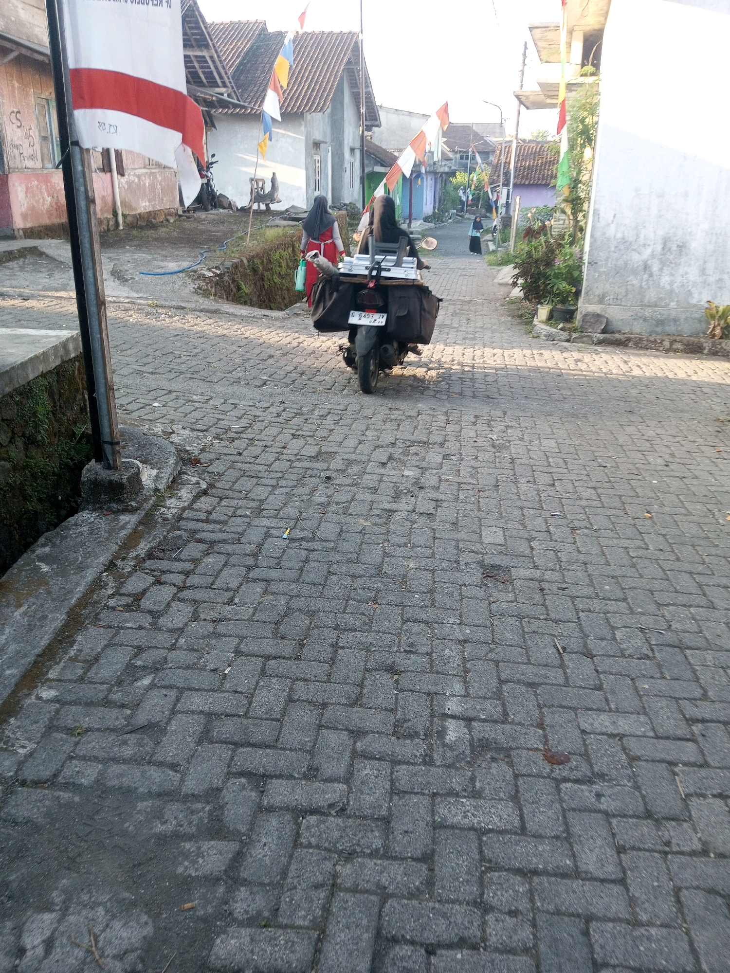 The image depicts a street scene in Andongsili, Indonesia, characterized by a paved road with interlocking brickwork. In the foreground, the road is lined with stone structures and vegetation along the left side. A flag with red and white stripes is prominently displayed on a pole on the left side of the image. A person wearing a black headscarf and a red and white outfit is walking away from the camera, holding a green container and a bag on their shoulder. They are walking towards a motorcycle parked on the road. The motorcycle, which appears to be a scooter, is loaded with bags and has a license plate with the inscription "G 6457 IV". Further down the street, other buildings and houses are visible, along with more flags. The environment appears to be a residential area with a somewhat narrow street. The overall lighting suggests it is daytime, possibly late afternoon due to the shadows.