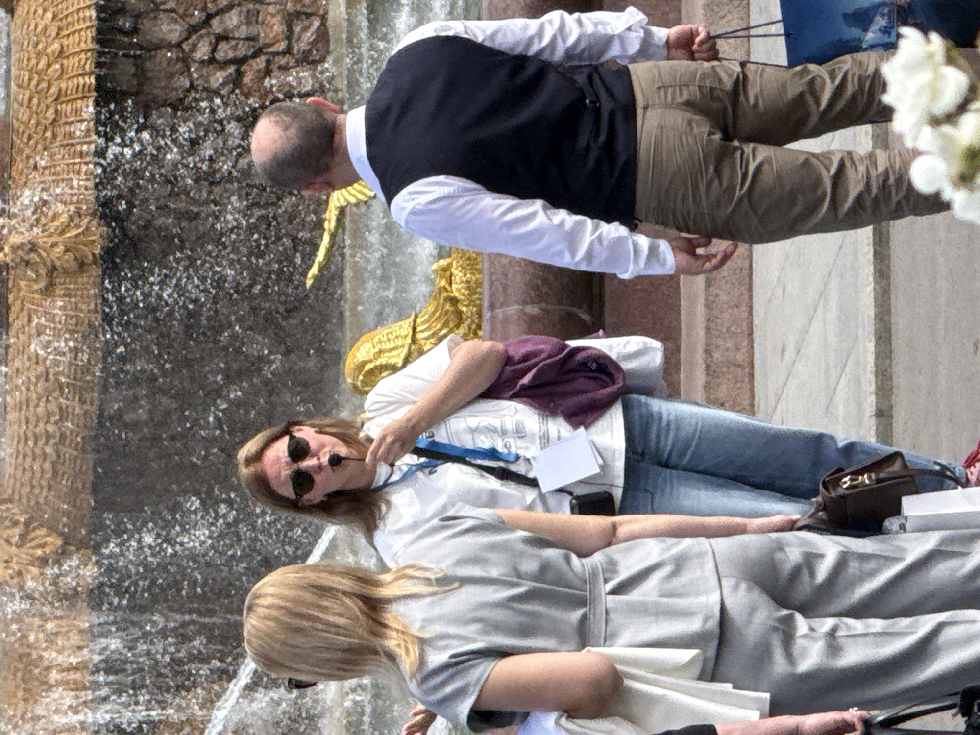 The image shows a group of people near a fountain, possibly in Moscow, Russia. The scene is shot from a high angle, looking down. In the foreground, a person in a light-colored jacket is seated, with their back to the camera. Next to them, another person is leaning forward, wearing a white shirt, dark vest, and tan pants. This person appears to be reaching towards the fountain. Further back, a person wearing sunglasses and a dark purple scarf is partially visible, leaning against something. The fountain itself is a prominent feature, with water splashing. The overall lighting suggests it is daytime. There is no visible text in the image.