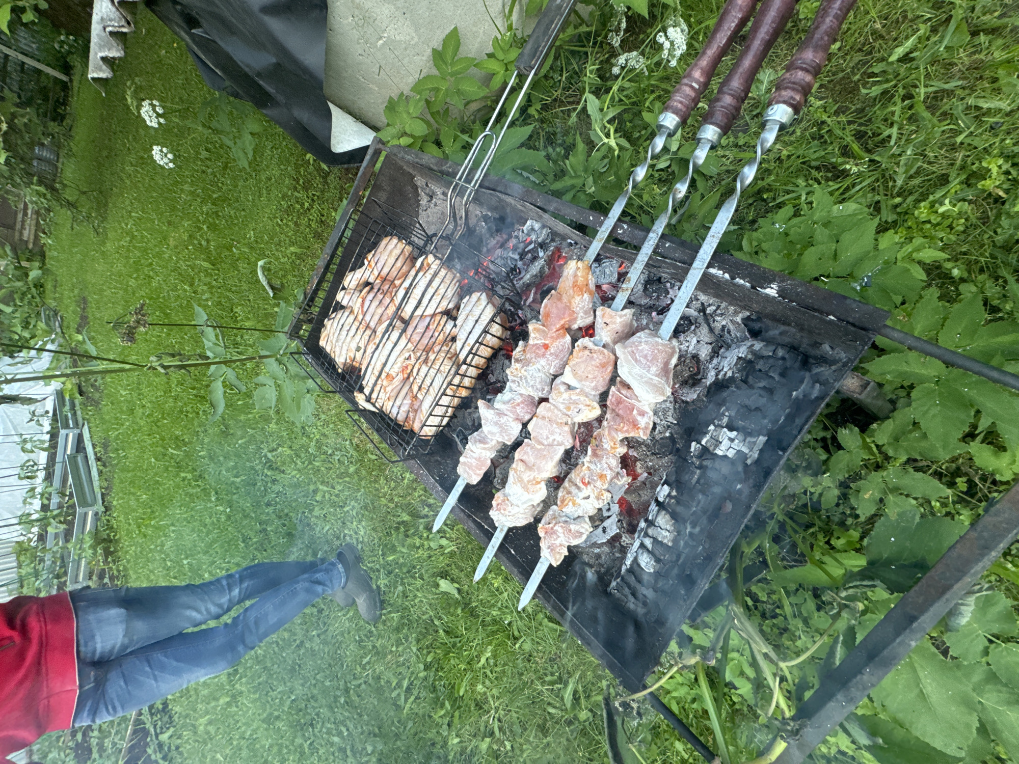 A top-down, eye-level shot captures a barbecue scene in a grassy yard in Stupino, Russia. The focus is on a black metal grill, filled with charcoal and glowing embers, supporting two cooking racks. On the lower rack, three metal skewers hold marinated meat, arranged in a row over the coals, with visible smoke rising. The upper rack contains a collection of raw chicken pieces, laid out in a grid pattern. To the left of the grill, a person's legs, clad in blue jeans and dark shoes, are visible, standing on the grass. The background features lush green grass, interspersed with plants and some white flowers. In the distance, a raised garden bed with plants is partially visible, suggesting a domestic outdoor setting. The overall ambiance is one of outdoor cooking and leisure.