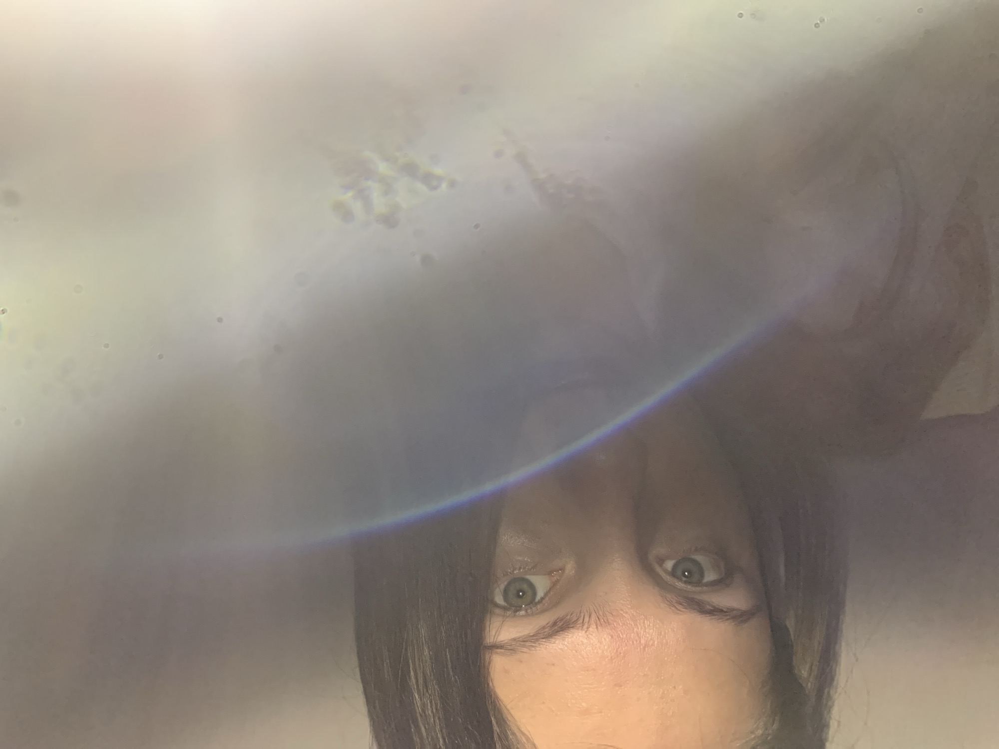 An inverted, close-up view of a person's face dominates the lower portion of the image. The subject's bright, alert green or hazel eyes are wide open, gazing directly forward. Dark hair frames the face, falling across the forehead and cheeks. The skin appears fair with some freckles visible on the forehead.
The upper and central parts of the image are dominated by a hazy, overexposed, and out-of-focus background, suggesting strong light sources. Scattered across this luminous haze are numerous small, circular, and irregularly shaped blurry spots, resembling dust particles or light artifacts. A prominent, arcing lens flare with a visible rainbow spectrum cuts across the image horizontally, just above the person's eyes, adding to the ethereal quality. The overall impression is one of an intimate, possibly spontaneous shot, with an unusual perspective. The location of the photo is Chaikovsky, Russia. There is no visible text in the image.