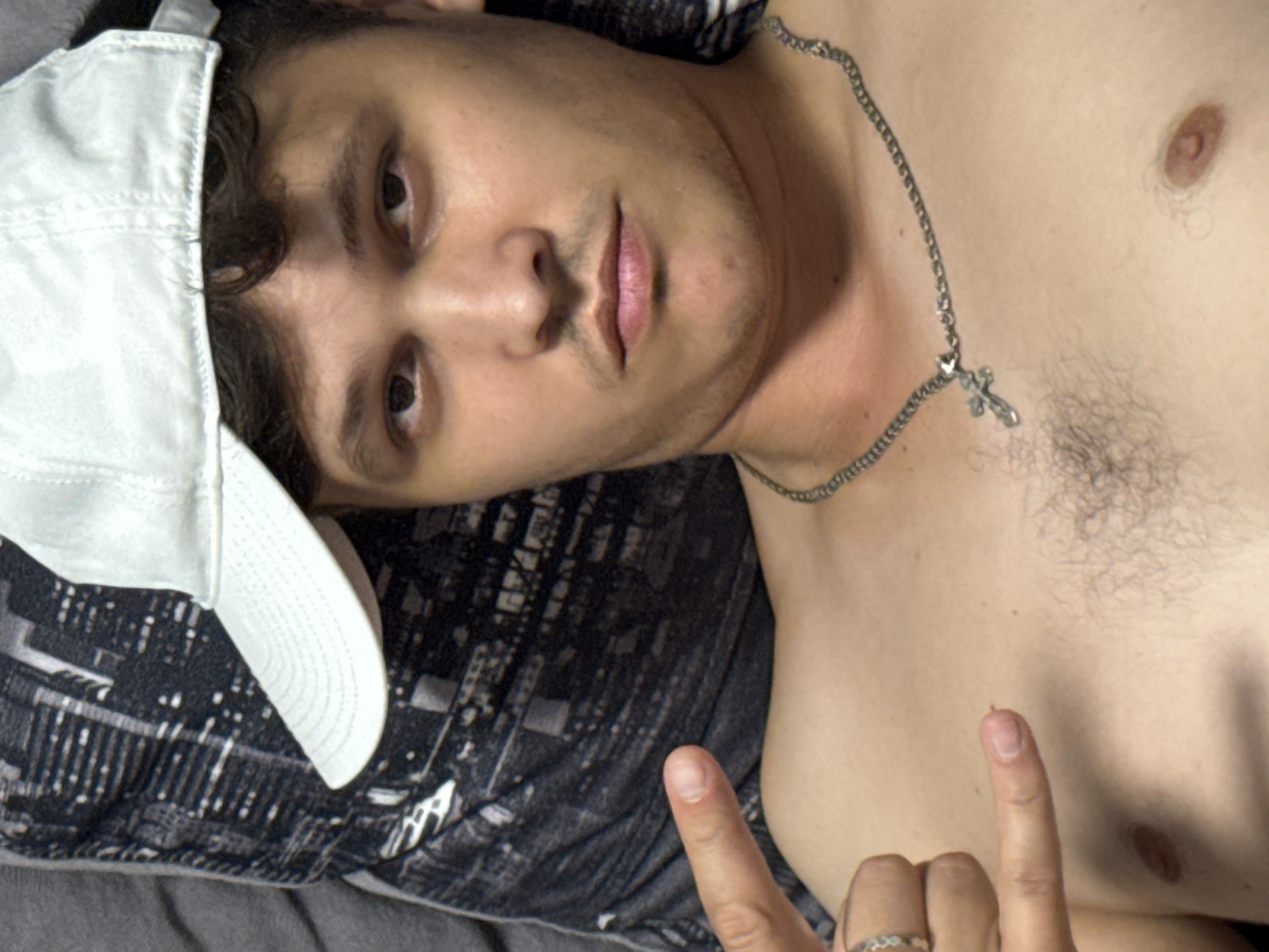 A partially clothed male lies on a patterned surface, possibly a bed or cushion. He wears a white baseball cap and a silver chain with a cross pendant. His fingers are visible in the lower part of the frame. The location is unknown.