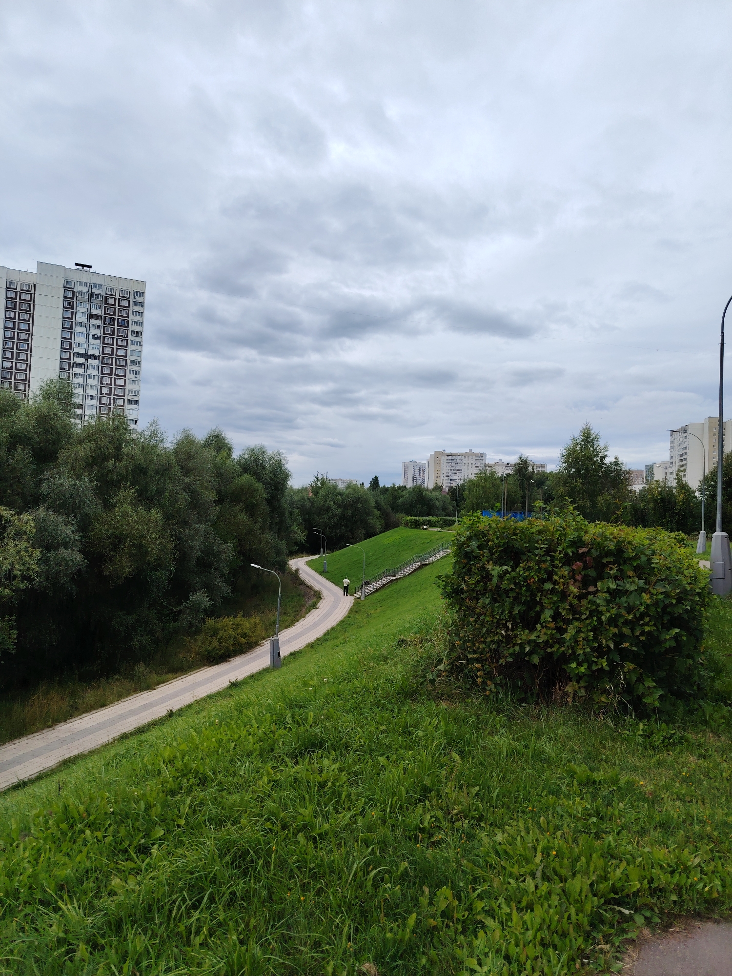 The image shows a curved pedestrian pathway winding through a grassy hillside with lush green trees on the left and right. Tall apartment buildings are visible in the background, consistent with an urban or suburban environment. A single person is walking on the pathway. The sky is overcast with clouds. The scene appears to be in the city of Zelenograd, Russia, specifically in the area of the 16th microdistrict, near the complex of buildings that includes "dom 1613" (1613 building).