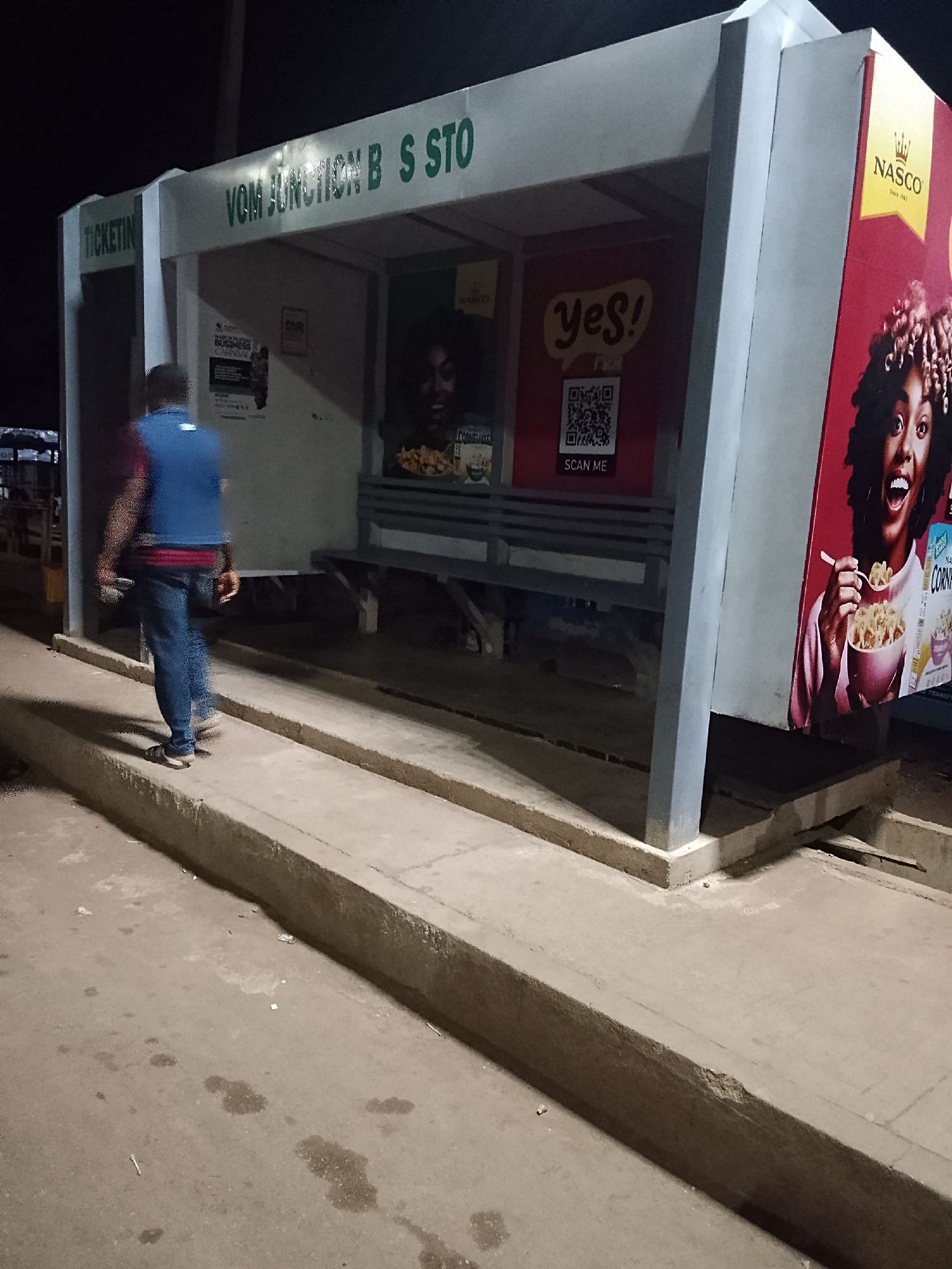 The image depicts an outdoor bus stop structure in Bukuru, Nigeria, indicated by the sign reading "VOM JUNCTION B S STO." The structure is constructed from gray metal supports and has a roof. Advertisements are affixed to the structure, including those for "NASCO" and a "yes!" campaign, featuring a QR code with the text "SCAN ME." A blurred individual wearing a blue vest and jeans is walking past the bus stop on a raised concrete platform. The area around the bus stop appears to be paved and dimly lit.