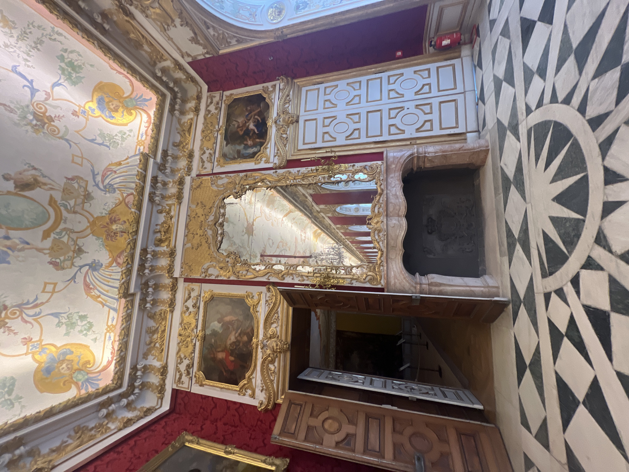 This image depicts the interior of a grand room, likely a hall or salon, within the Neues Schloss (New Palace) in Oberschleißheim, Germany. The room is opulently decorated with gilded moldings, ornate paintings on the ceiling, and elaborate tapestries or wallpaper in a deep red hue.
A large, intricately framed mirror dominates the center of the room, reflecting a long, chandelier-lit corridor. To the left of the mirror, a fireplace with a marble surround is visible, featuring a dark grate with decorative carvings, possibly a coat of arms. To the right of the mirror, a tall, white double door with gold trim and oval insets is set into the wall, contrasting with the red surroundings.
In the foreground, the floor is covered with a striking geometric pattern of black and white diamond-shaped tiles, with a circular motif in the lower right quadrant featuring a star-like design. Several doorways are visible, with one set of double wooden doors on the left standing ajar, revealing a glimpse into another, darker space.
The overall impression is one of historical grandeur and opulent Baroque or Rococo architecture and interior design. There are no people visible in the frame. A red fire extinguisher is mounted on the wall to the right of the white double doors.
