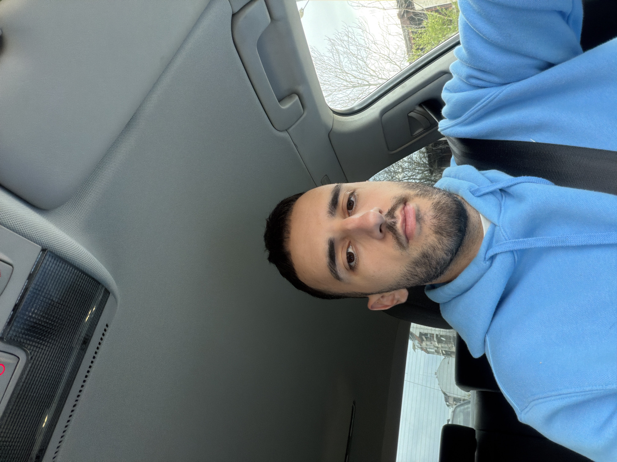 A man with dark hair and a beard, wearing a light blue hoodie, is seated in the driver's seat of a car. The car appears to have a sunroof open. The interior of the car is gray. The image's orientation is turned sideways. Outside the window, buildings and trees are visible. Based on the architecture visible through the window, the location cannot be confirmed as Plovdiv, Bulgaria.