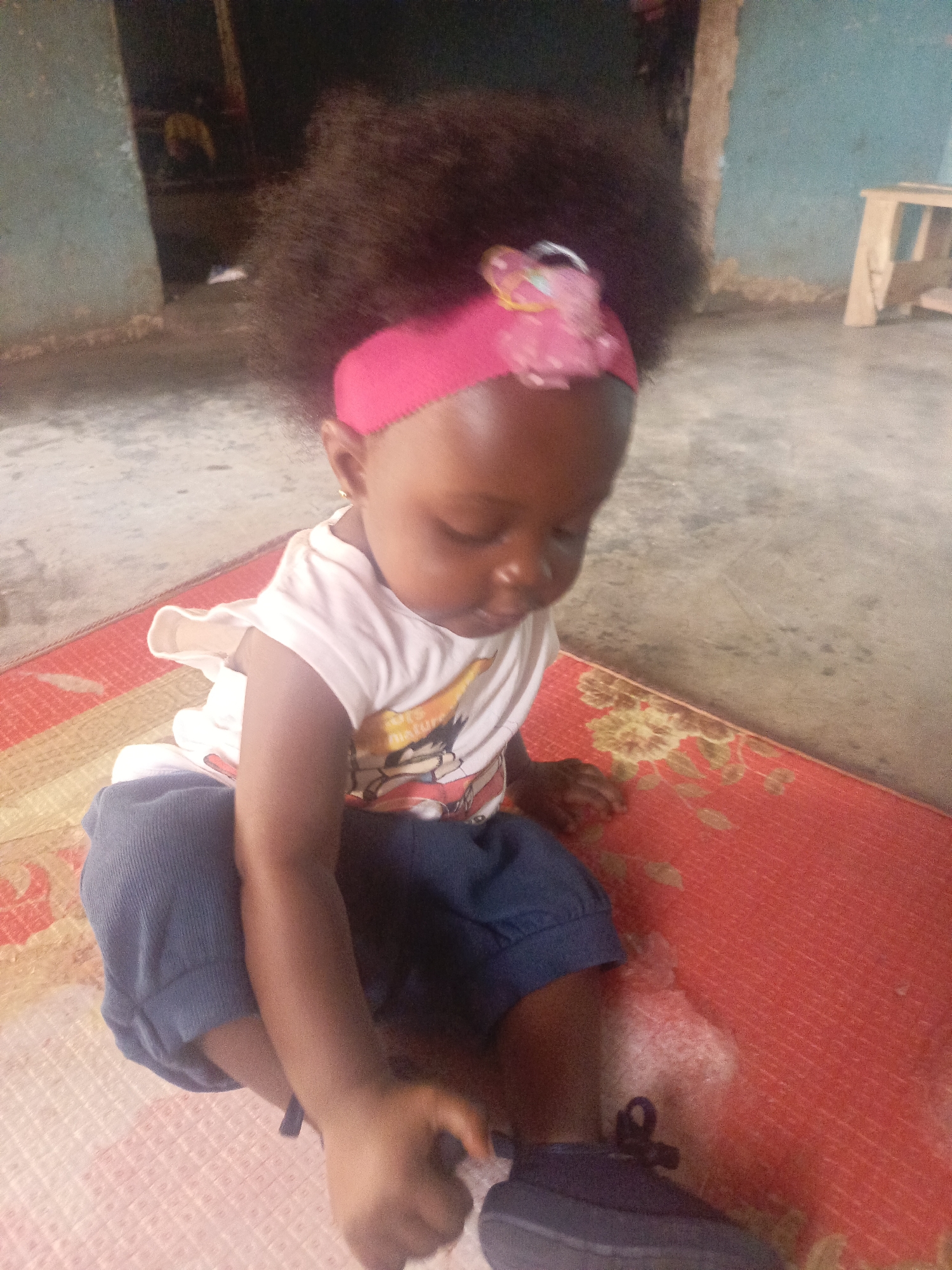 A young child is seated on a red and yellow patterned mat inside a building in an unknown city. The child is wearing a white t-shirt with a cartoon graphic, blue pants, and a pink headband with a pink decorative object. The child is reaching down to adjust or touch a blue shoe. The building's interior has faded blue walls and a gray concrete floor. A small wooden stool is visible in the background.