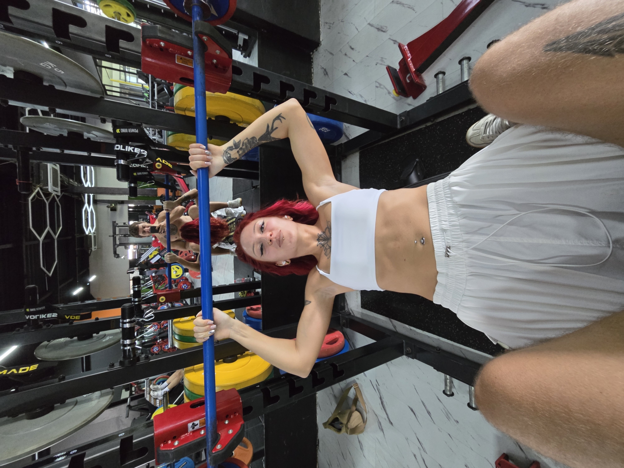 The image is taken in a gym in Unknown, Unknown. A person with red hair, wearing a white sports bra and white shorts, is holding a blue exercise bar. They have multiple tattoos on their arms and chest. There is gym equipment visible in the background, including weight plates on a rack, and mirrored surfaces that reflect the scene. Another person's legs and feet, wearing white shoes and white shorts, are partially visible in the foreground. The floor is tiled with a marble-like pattern.