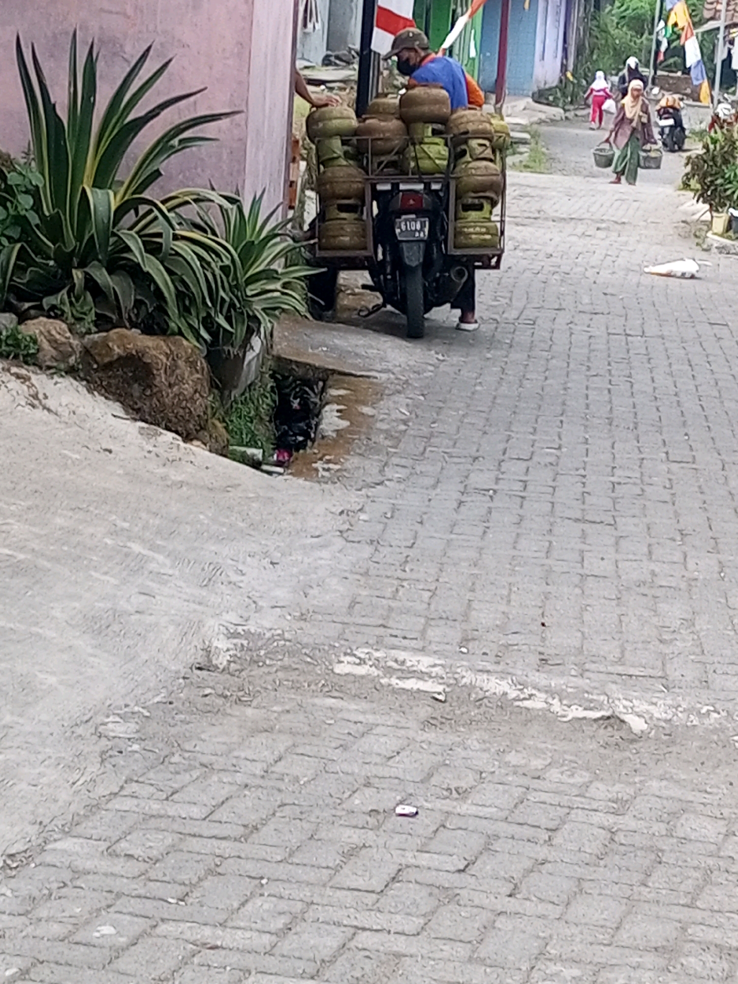 A man is seen on a motorcycle in Andongsili, Indonesia, transporting gas cylinders. The motorcycle is equipped with a rack holding multiple gas cylinders. The man is wearing a blue shirt, an orange undershirt, a dark baseball cap, and a face mask. He is facing away from the camera, appearing to be in the process of moving or delivering the gas. In the background, two women are walking on a cobblestone street, each carrying a basket. One woman wears a light-colored hijab and a dark top with green pants, while the other wears a red top and white hijab. The street is paved with gray cobblestones. A large green plant with pointed leaves is visible on the left side of the image, next to a light pink wall. The environment appears to be a residential or commercial area with buildings visible in the distance.